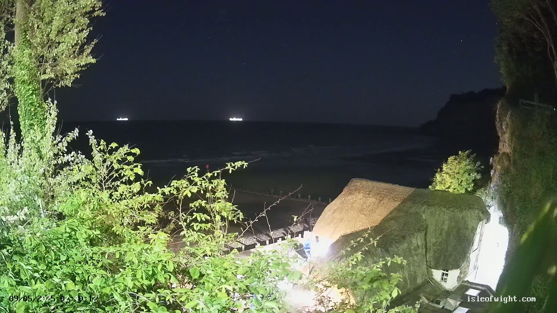 At night, a thatched-roof building sits on a cliff overlooking a dark ocean with two distant lights, possibly ships, and some vegetation in the foreground.