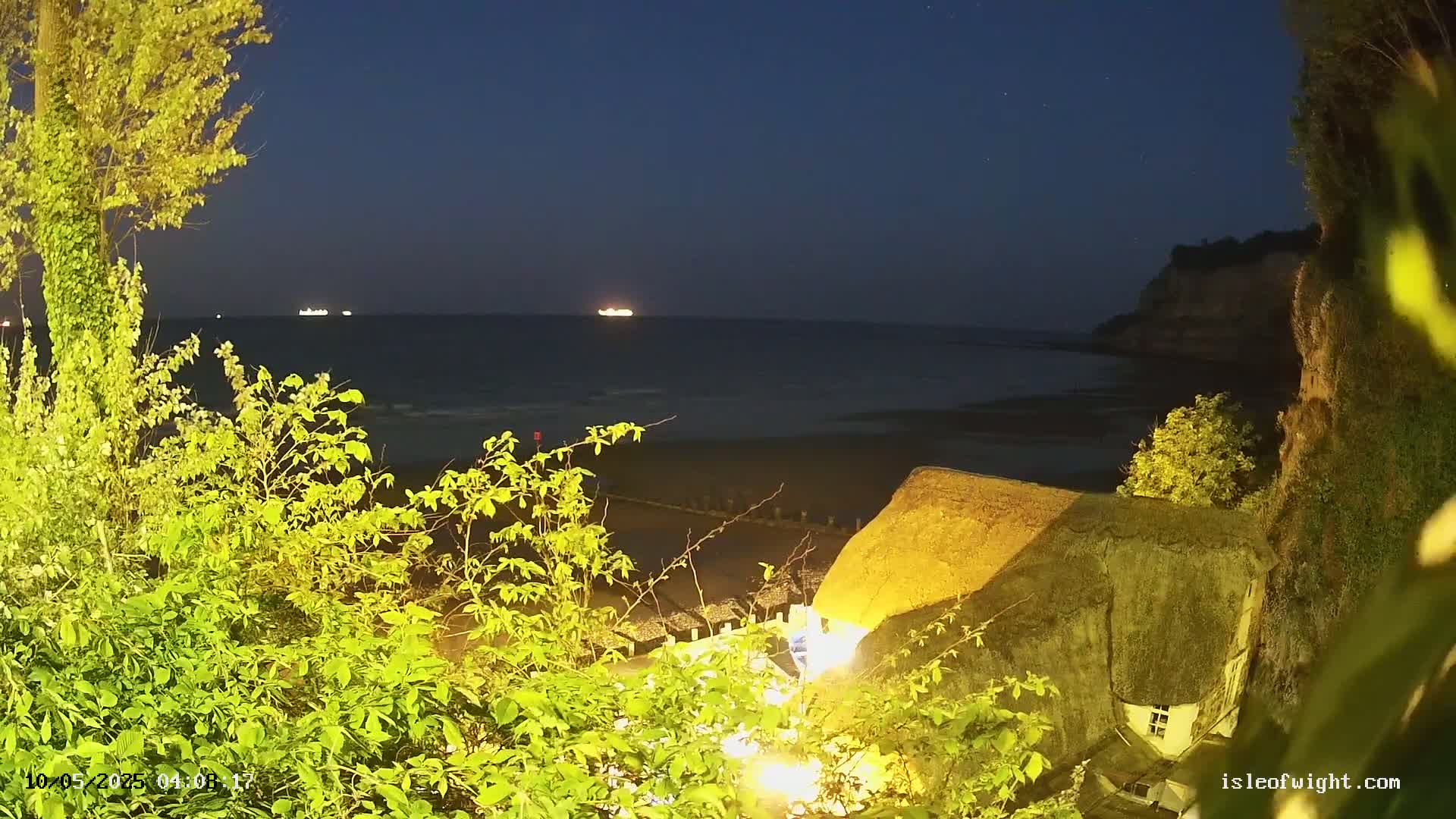 A thatched-roof cottage is illuminated at night overlooking a calm, dark sea with a few distant lights from ships, under a mostly clear night sky.