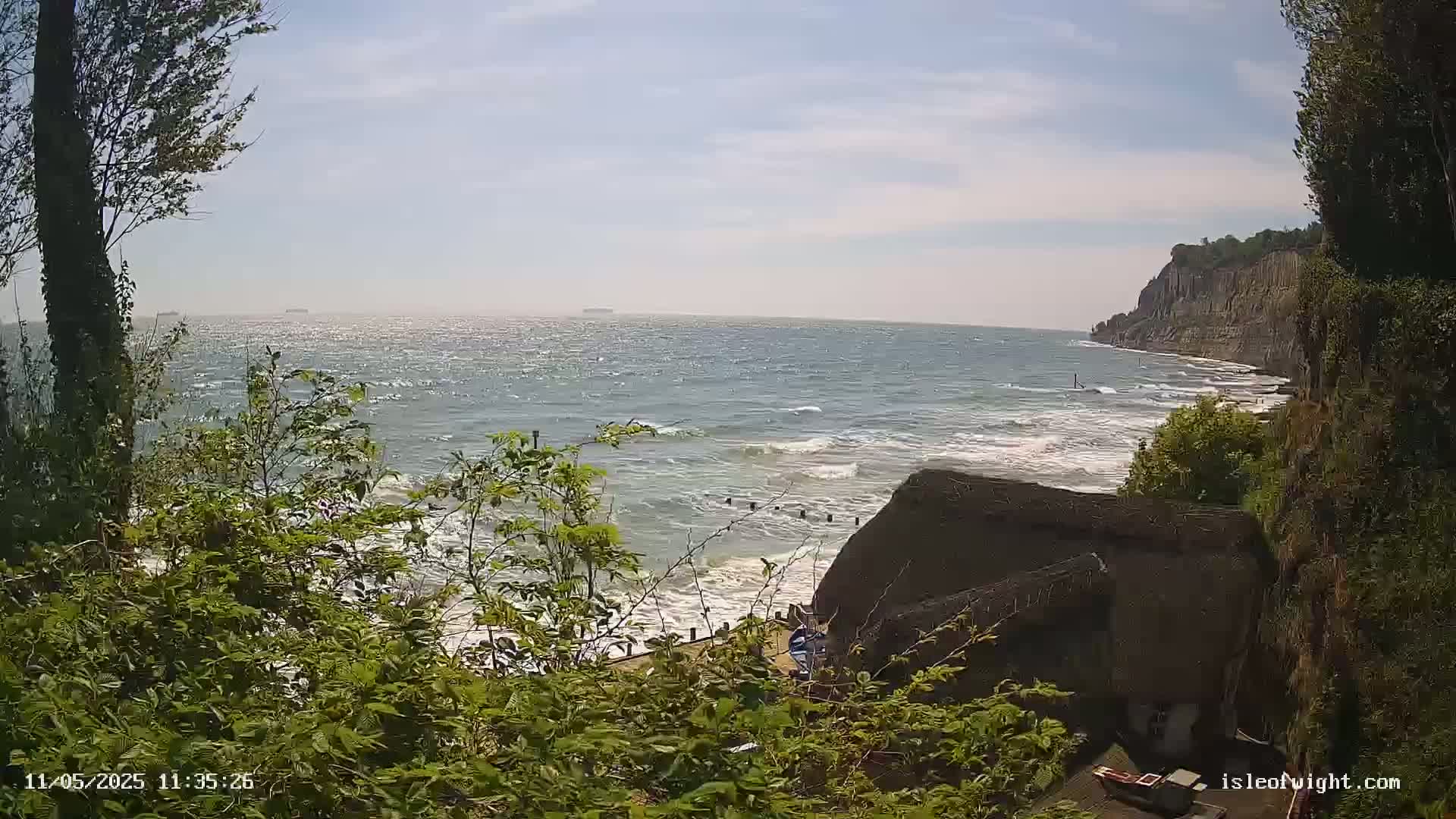 A windy, sunny day overlooks a rocky coastline with a thatched roof building partially visible, and a choppy sea with small waves.
