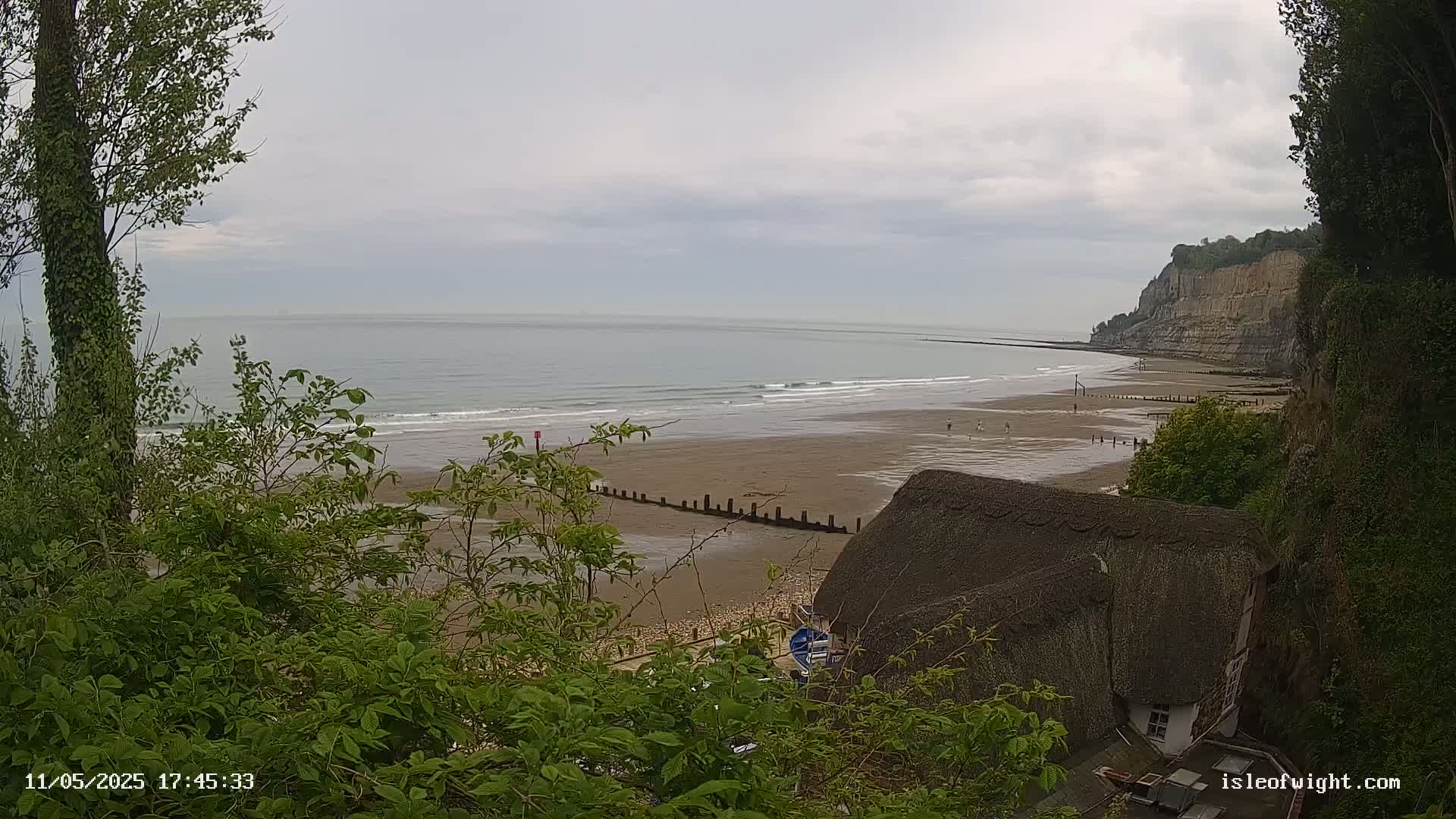 Overlooking a sandy beach with gentle waves under a cloudy sky, a thatched-roof house is partially visible nestled among lush green vegetation on a cliffside.