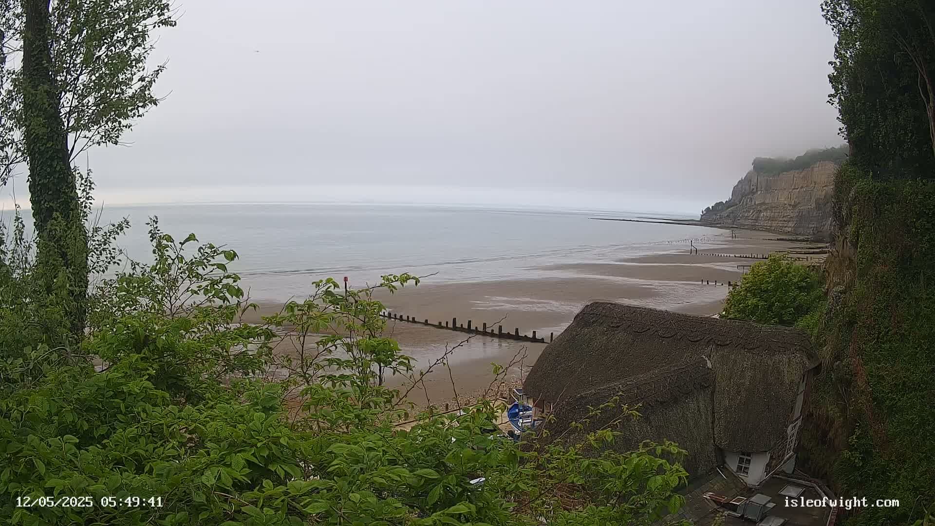 A calm, overcast day reveals a sandy beach stretching to a low cliff, partially obscured by vegetation in the foreground, and a thatched-roof building nestled at the base of the cliff.
