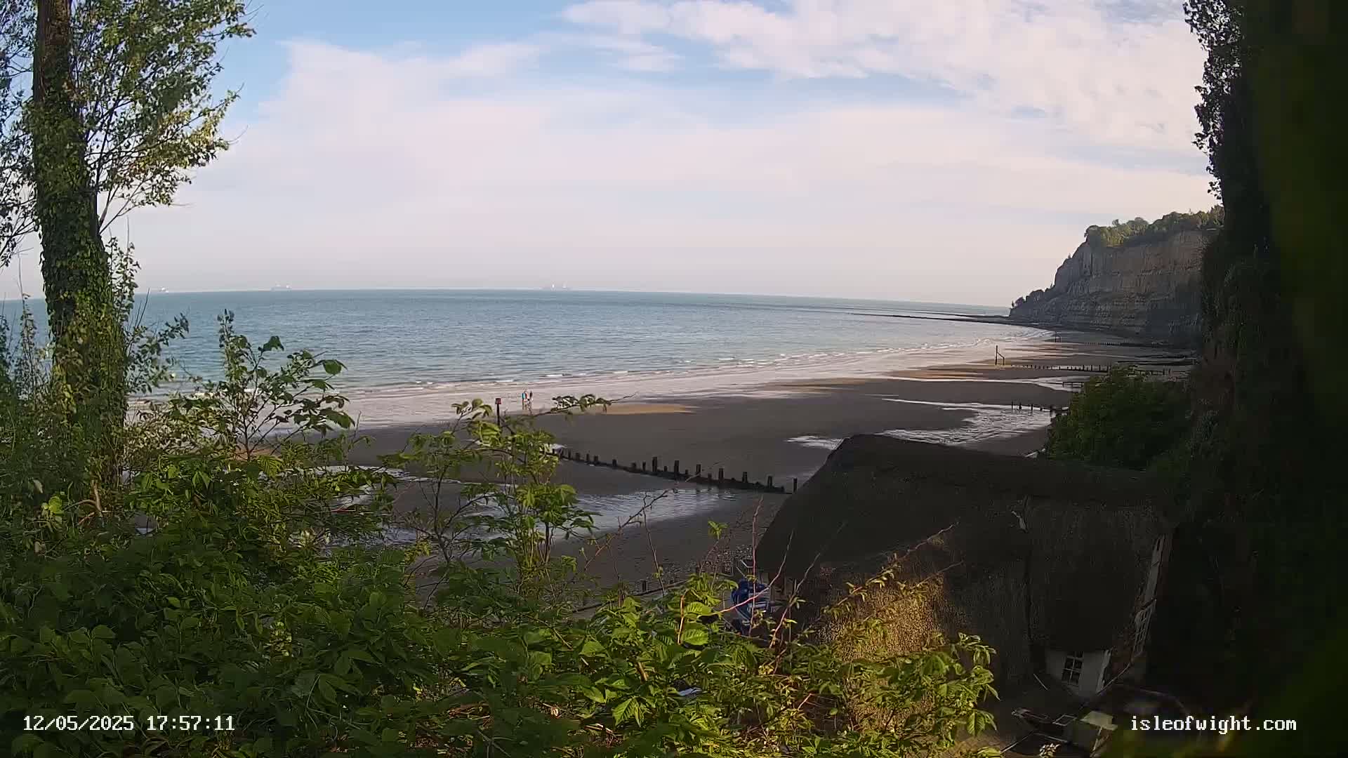 A sandy beach stretches out to a calm sea under a partly cloudy sky, viewed from behind leafy vegetation and a thatched roof building nestled against a cliff.