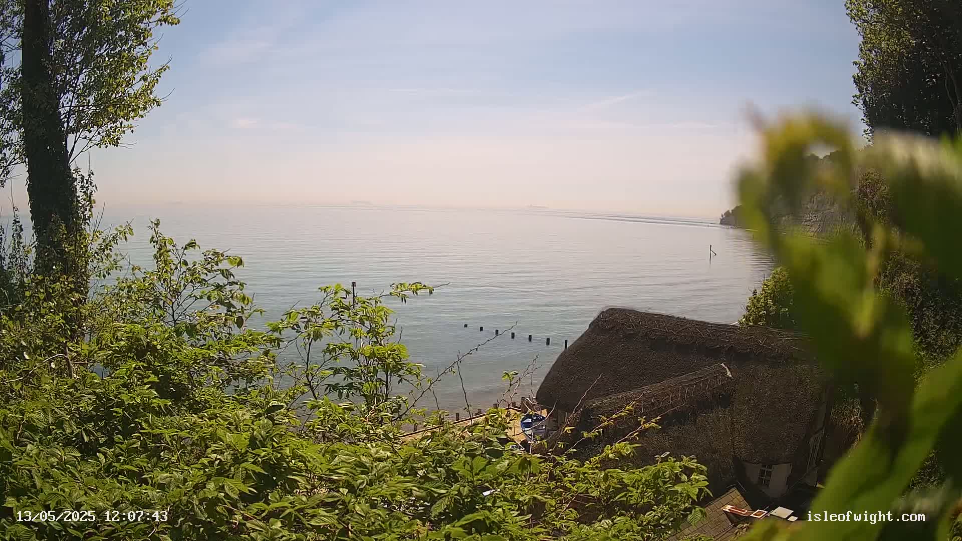 A calm, hazy sea is visible from an elevated vantage point partially obscured by lush green foliage, with a thatched-roof building nestled below.