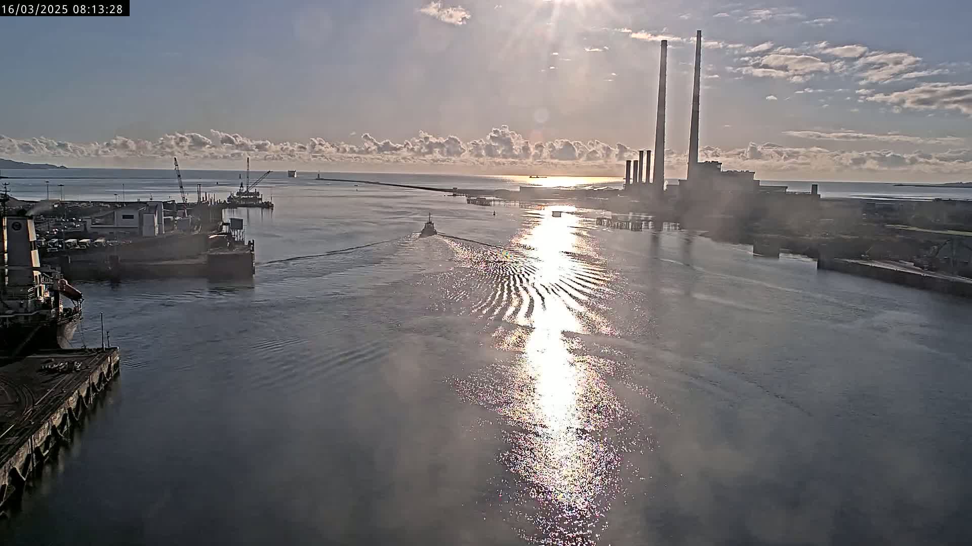 A tugboat travels through a calm waterway toward a power plant on a sunny day with scattered clouds.