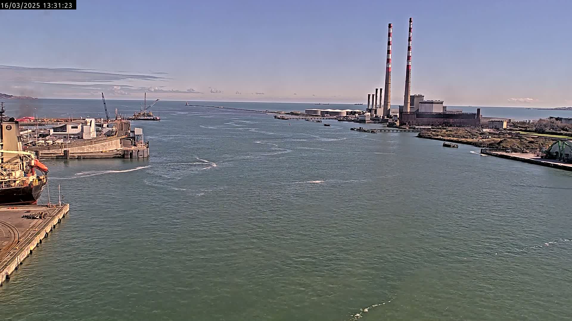 A large cargo ship is docked at a port, with a power plant and its tall smokestacks visible in the distance across a calm body of water under a mostly clear sky.