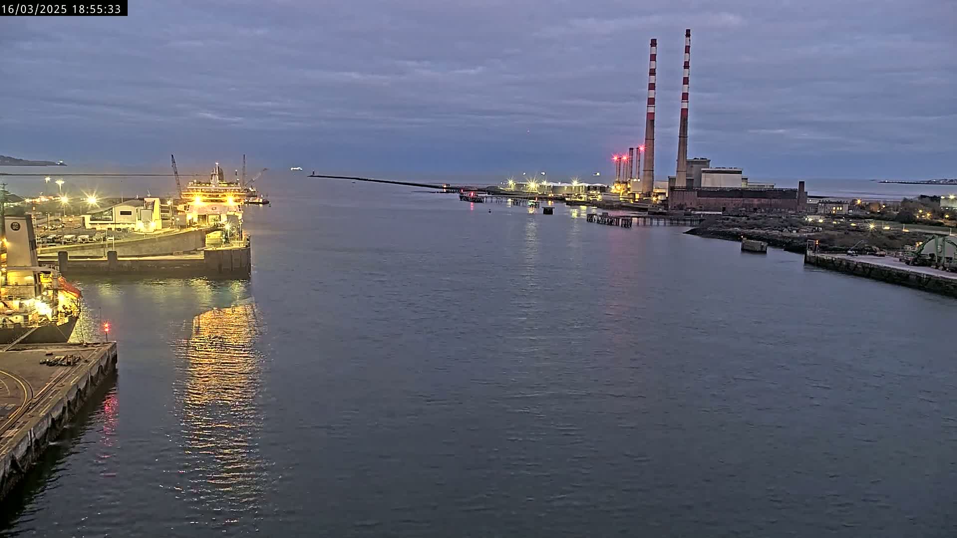 A twilight view of a harbor with docked ships, a power plant with tall chimneys, and a breakwater extending into calm water.