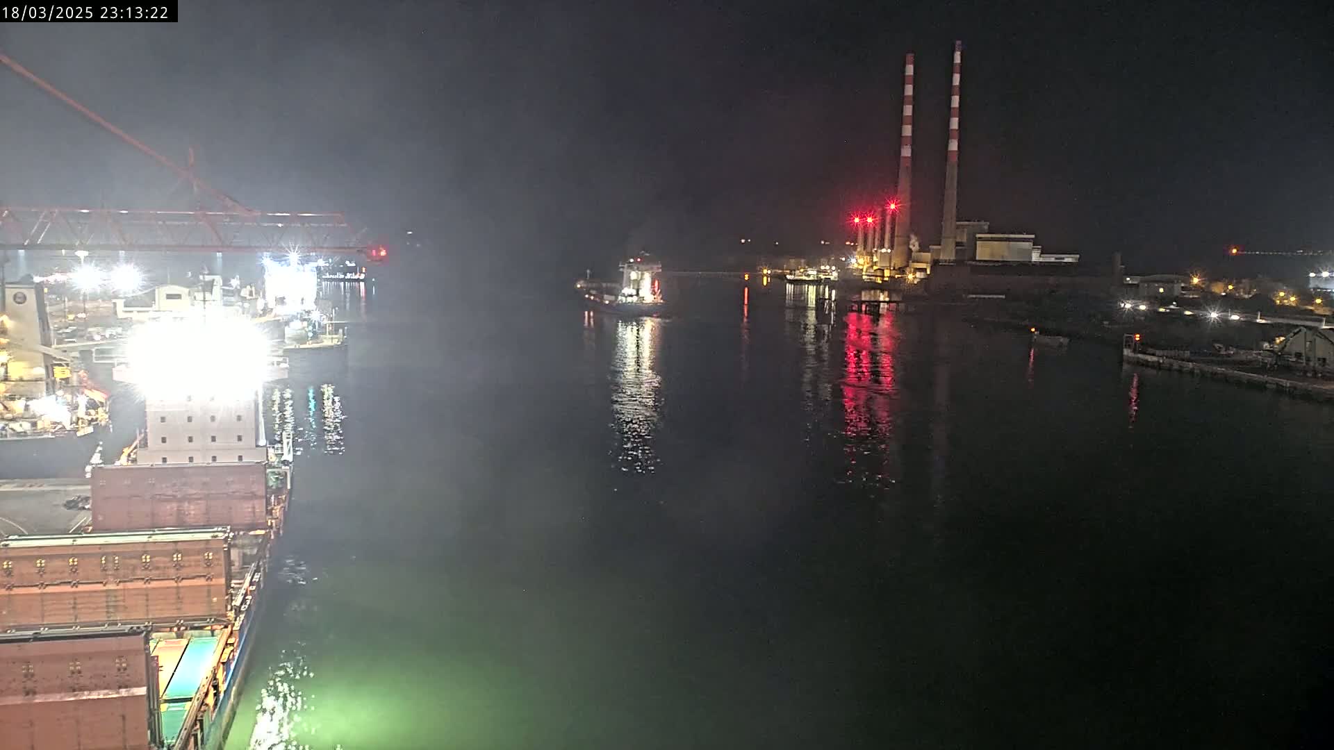 A nighttime view of a harbor shows a large cargo ship docked, another ship passing by in the water, and a power plant with tall smokestacks in the background.