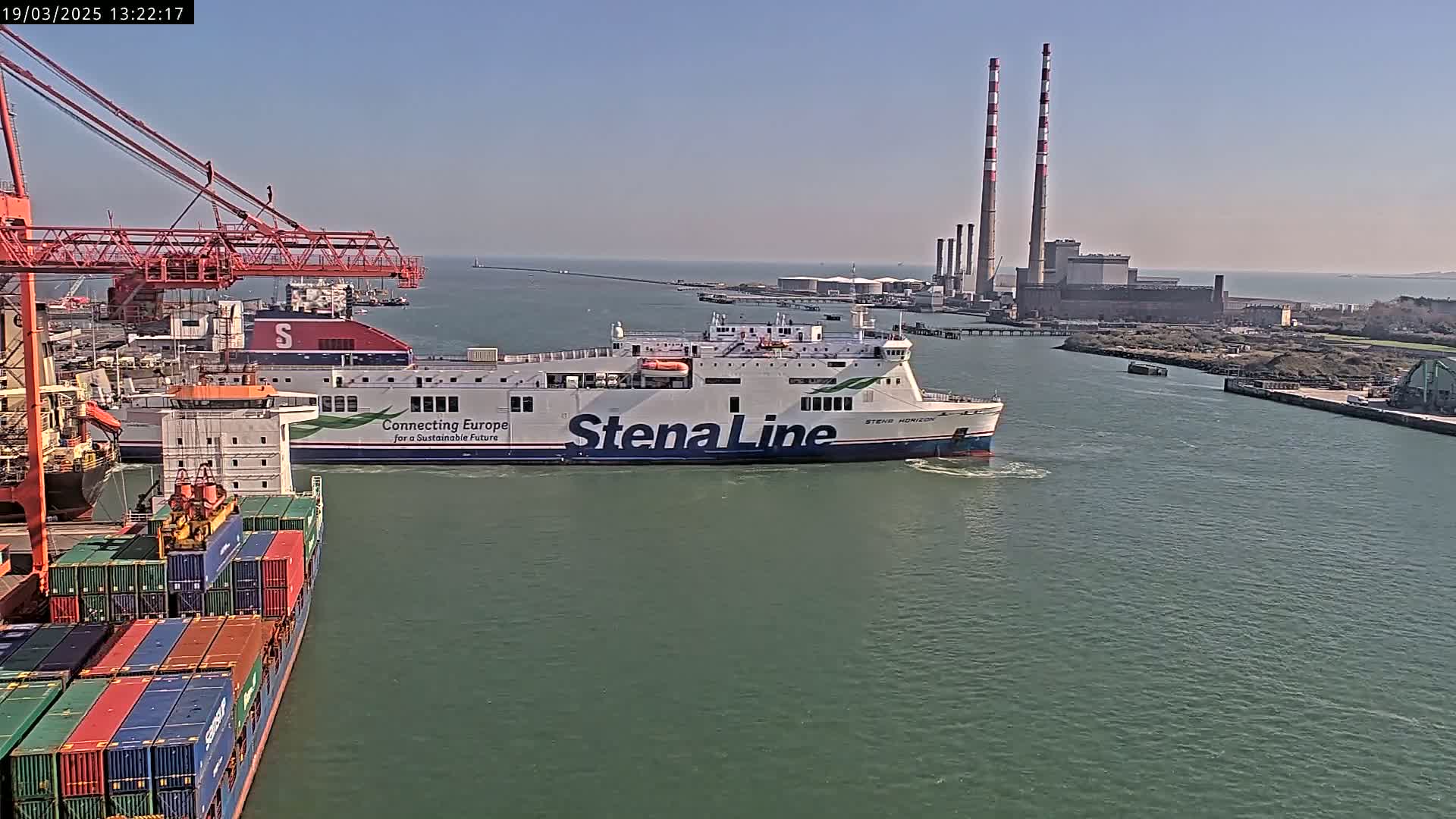 A large ferry passes by a container ship and industrial buildings under a clear, sunny sky.