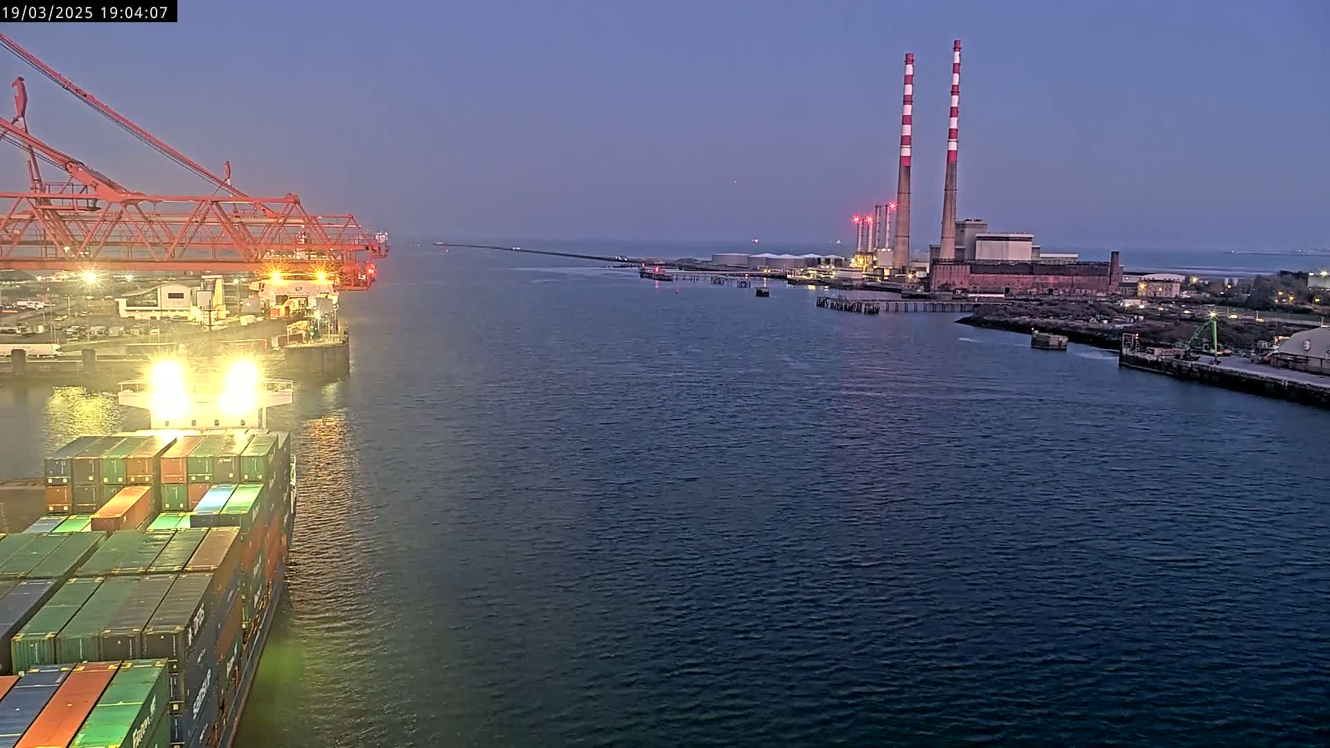 A container ship sails past a port at dusk, with large industrial structures and tall chimneys visible in the distance.