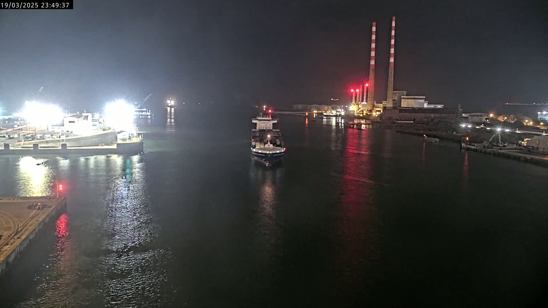 A cargo ship sails through a dark, calm waterway at night past an industrial facility with tall smokestacks.