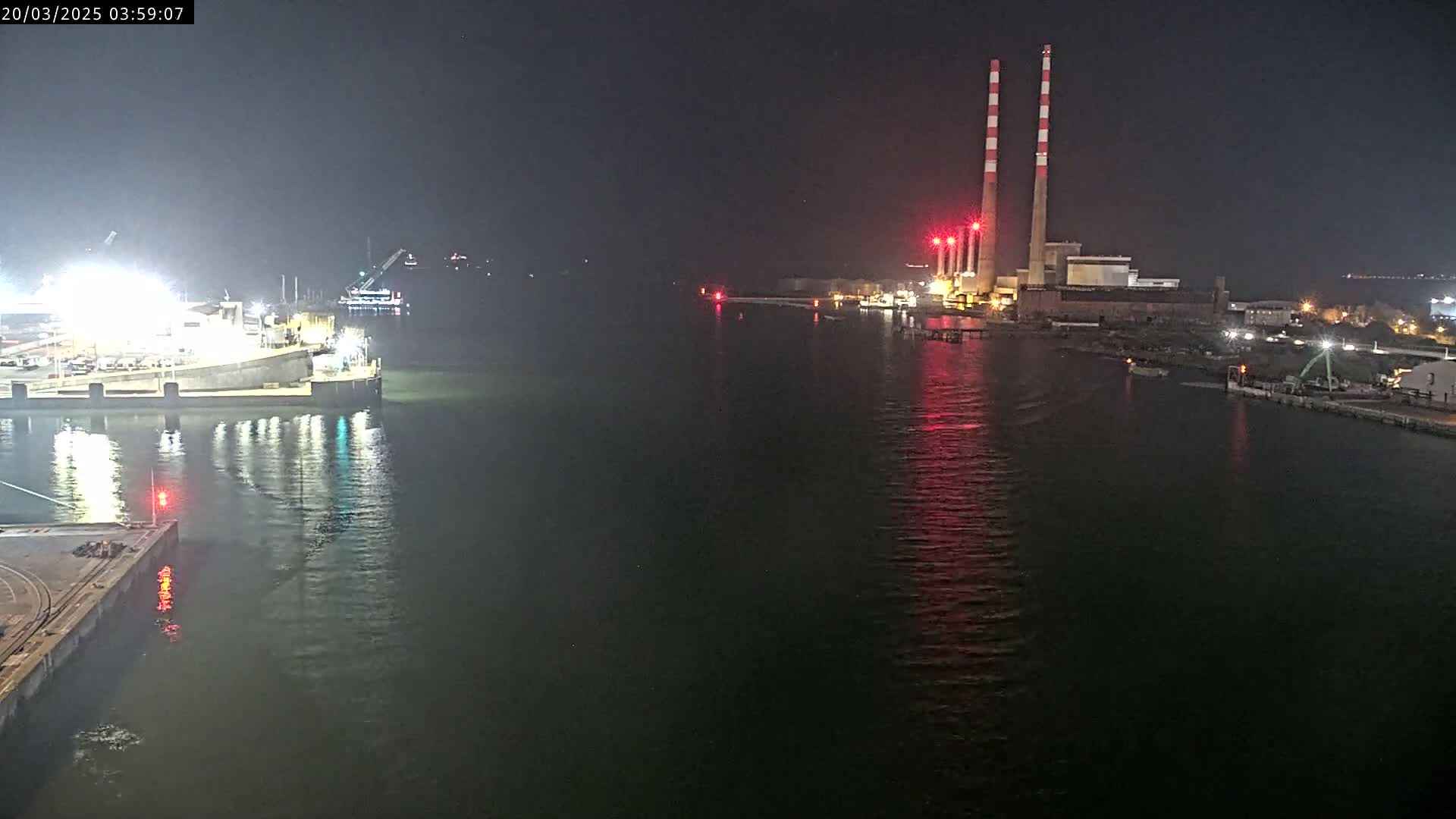 A nighttime view of a harbor with a large industrial facility and several docked ships, reflecting lights on calm water.