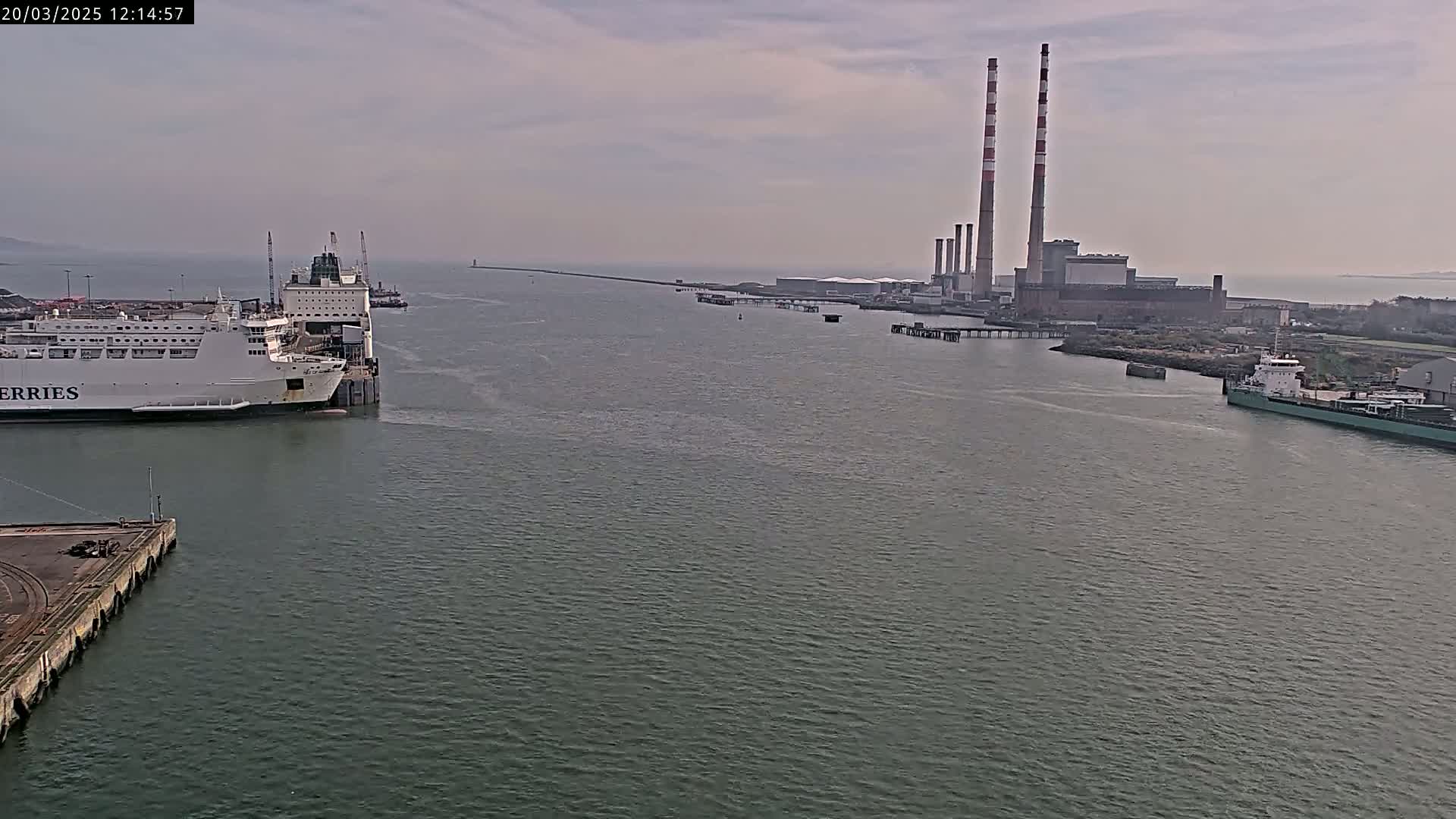 A ferry is docked at a port, with a power plant and other ships visible in the distance under an overcast sky.