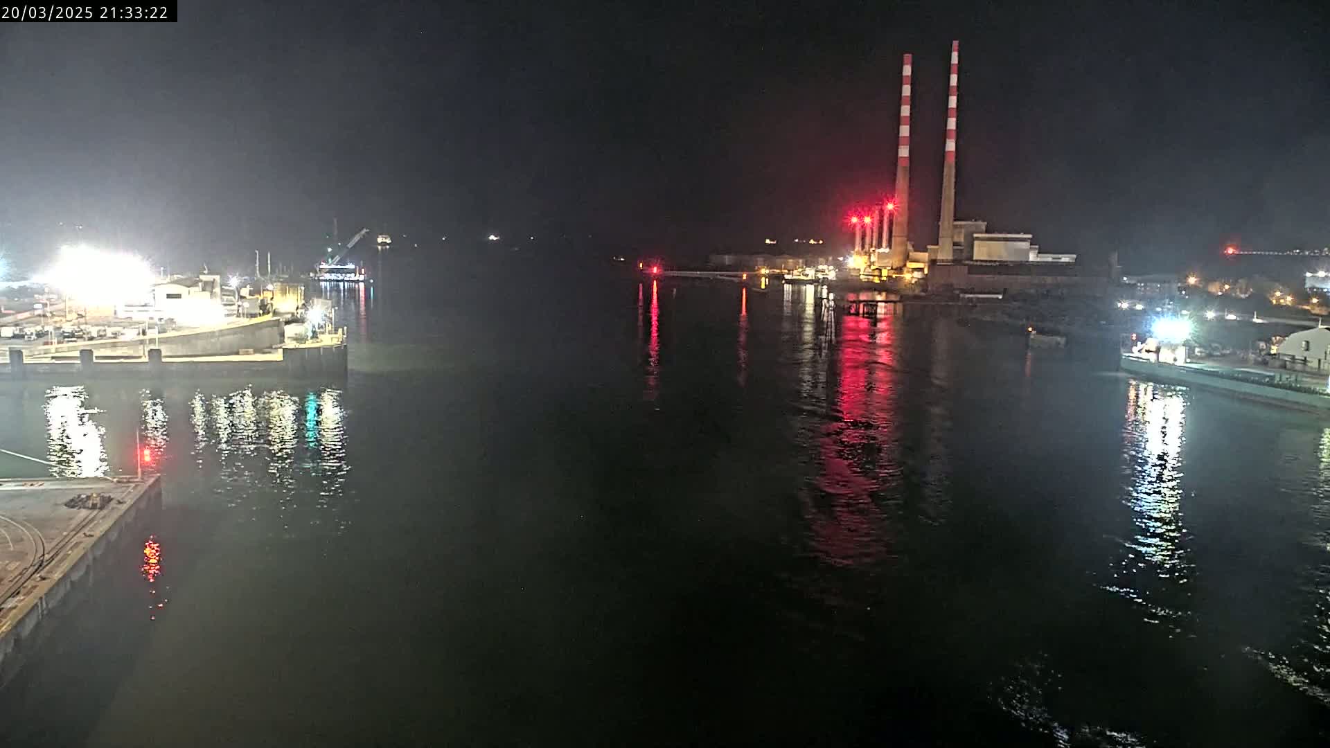 At night, a dark body of water reflects the lights of a dock, a distant industrial plant with tall smokestacks, and a ship.