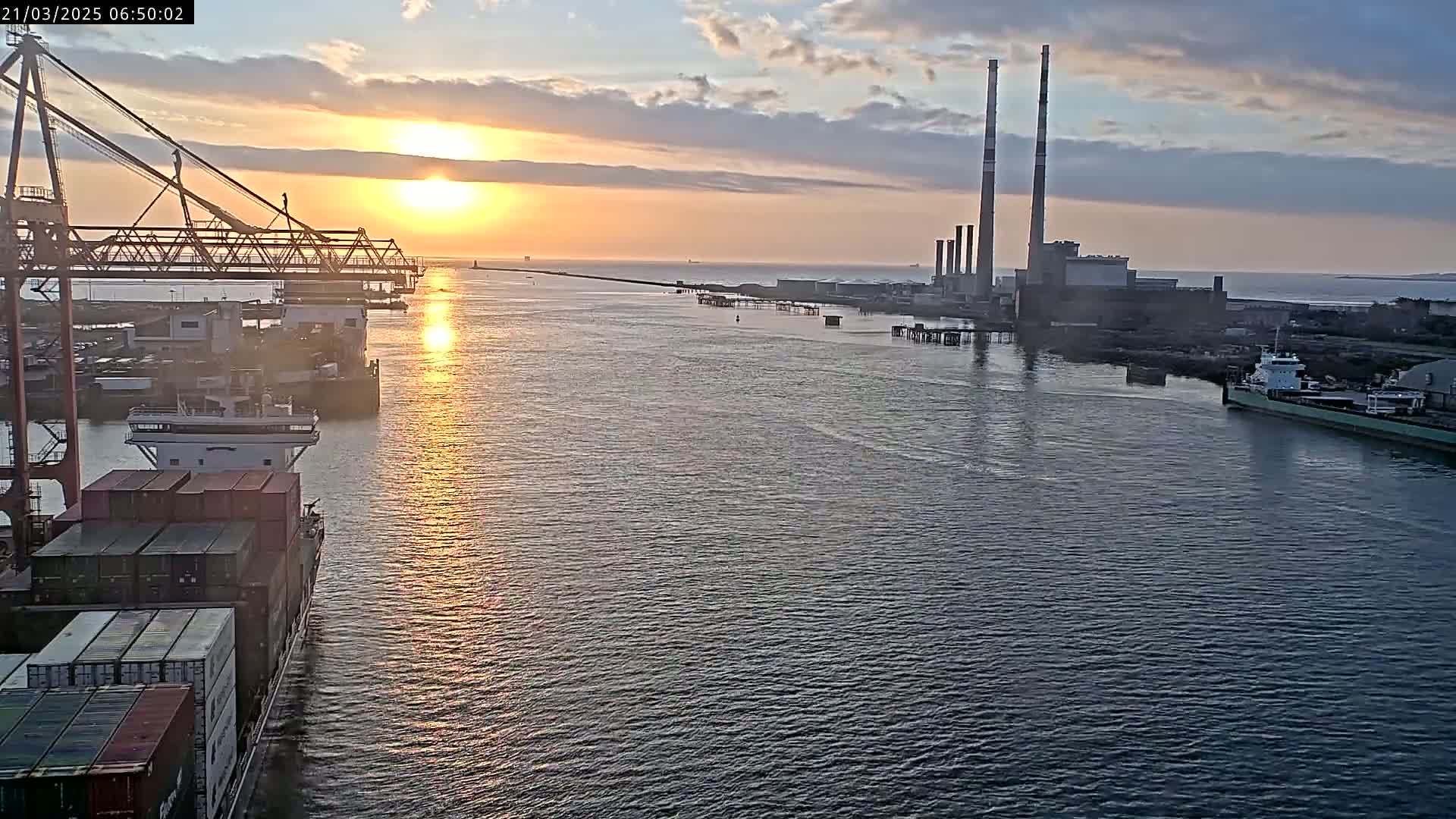 A container ship sails past a port at sunrise under partly cloudy skies, with a large industrial facility visible in the distance.
