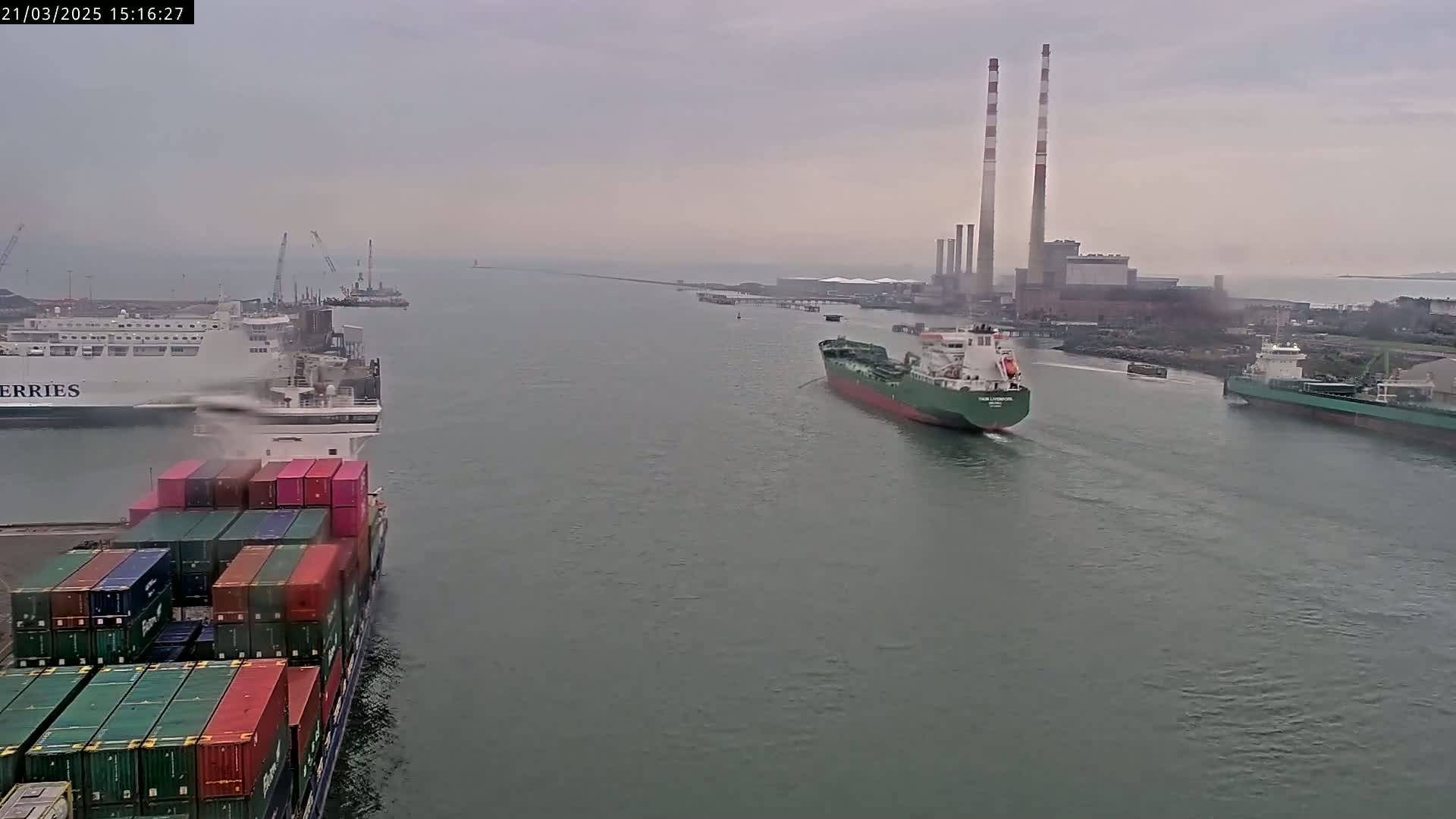 Several ships, including a container ship and a tanker, navigate a waterway on an overcast day, with a large industrial facility visible in the background.
