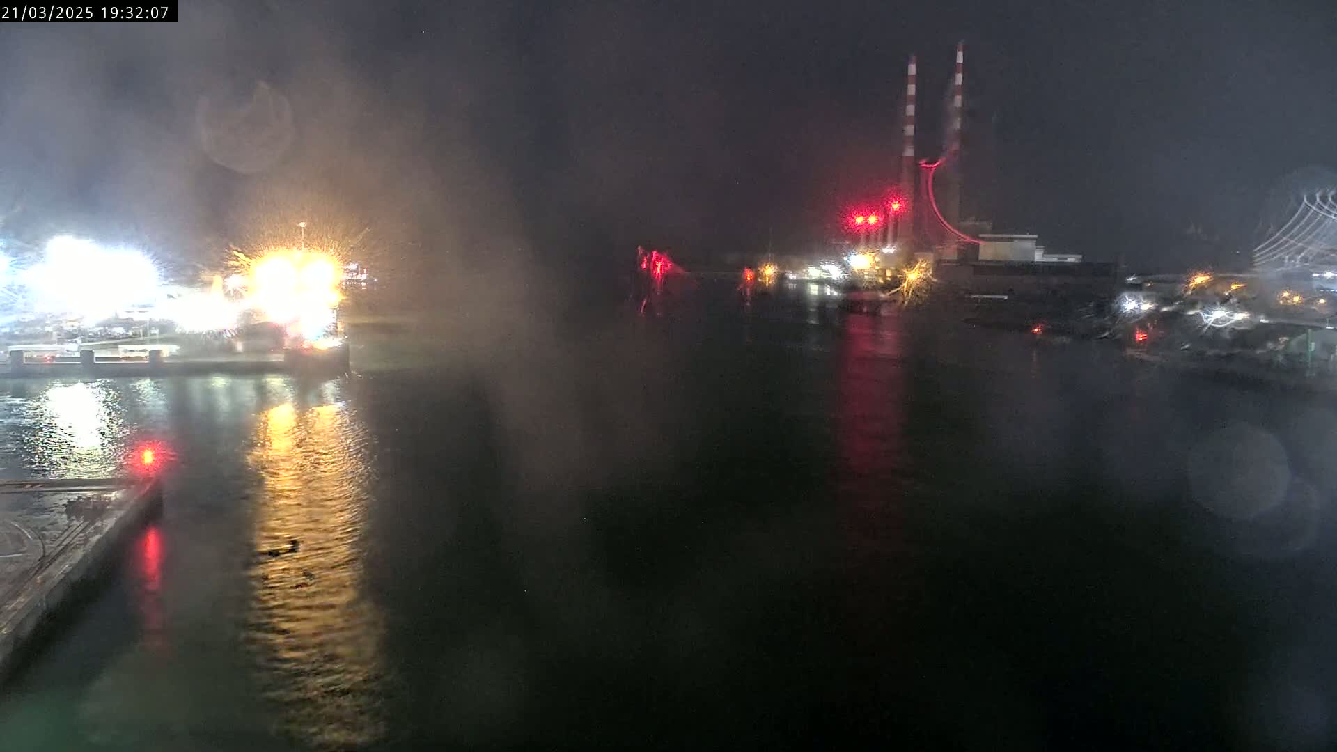 A nighttime view of a harbor with brightly lit industrial structures across the water, viewed through rain-streaked glass.