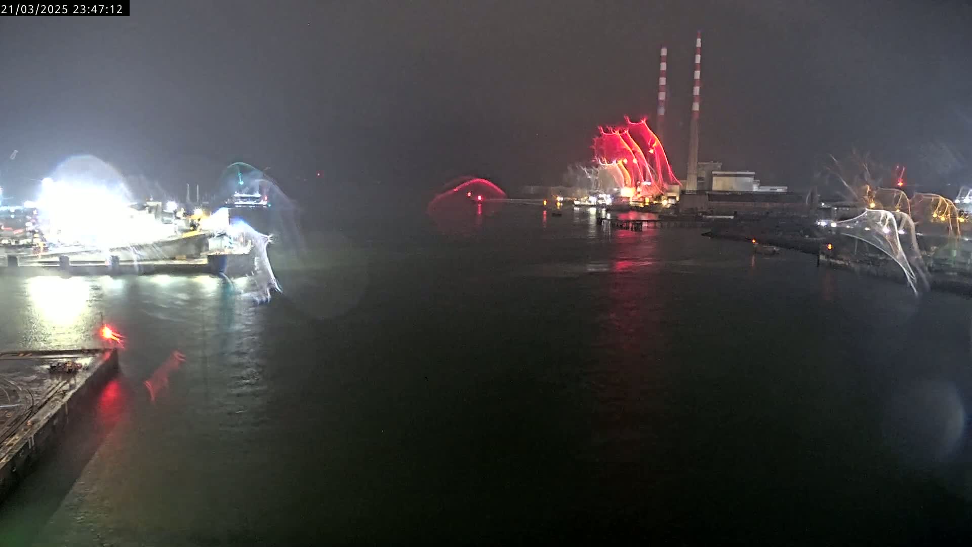 At night, in dark and seemingly wet conditions, several boats are illuminated by bright white and red lights near a large industrial structure with tall smokestacks.