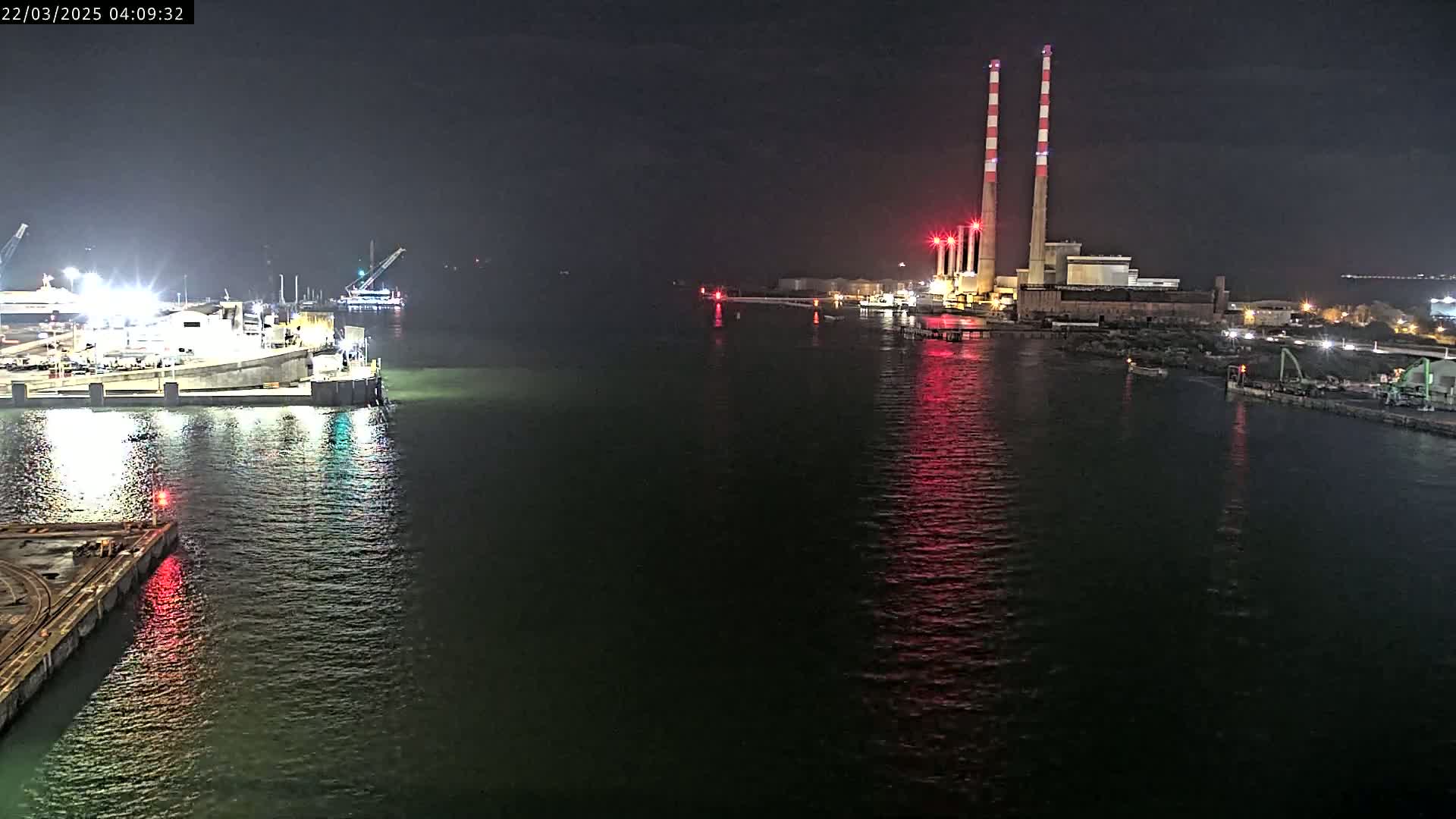 A nighttime view of a calm body of water with a brightly lit industrial facility and docked vessels on either side.