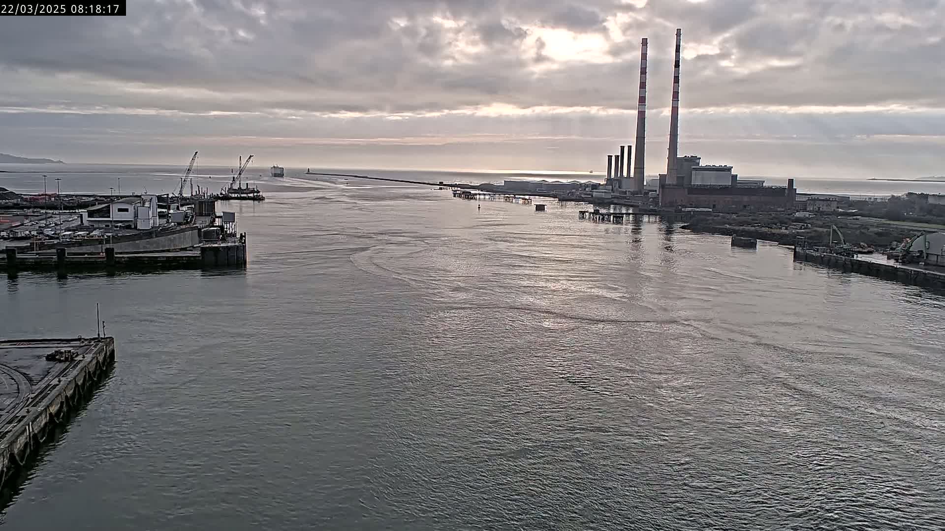 A wide, calm waterway shows a port with docked ships and a large industrial plant with tall smokestacks under a cloudy sky.
