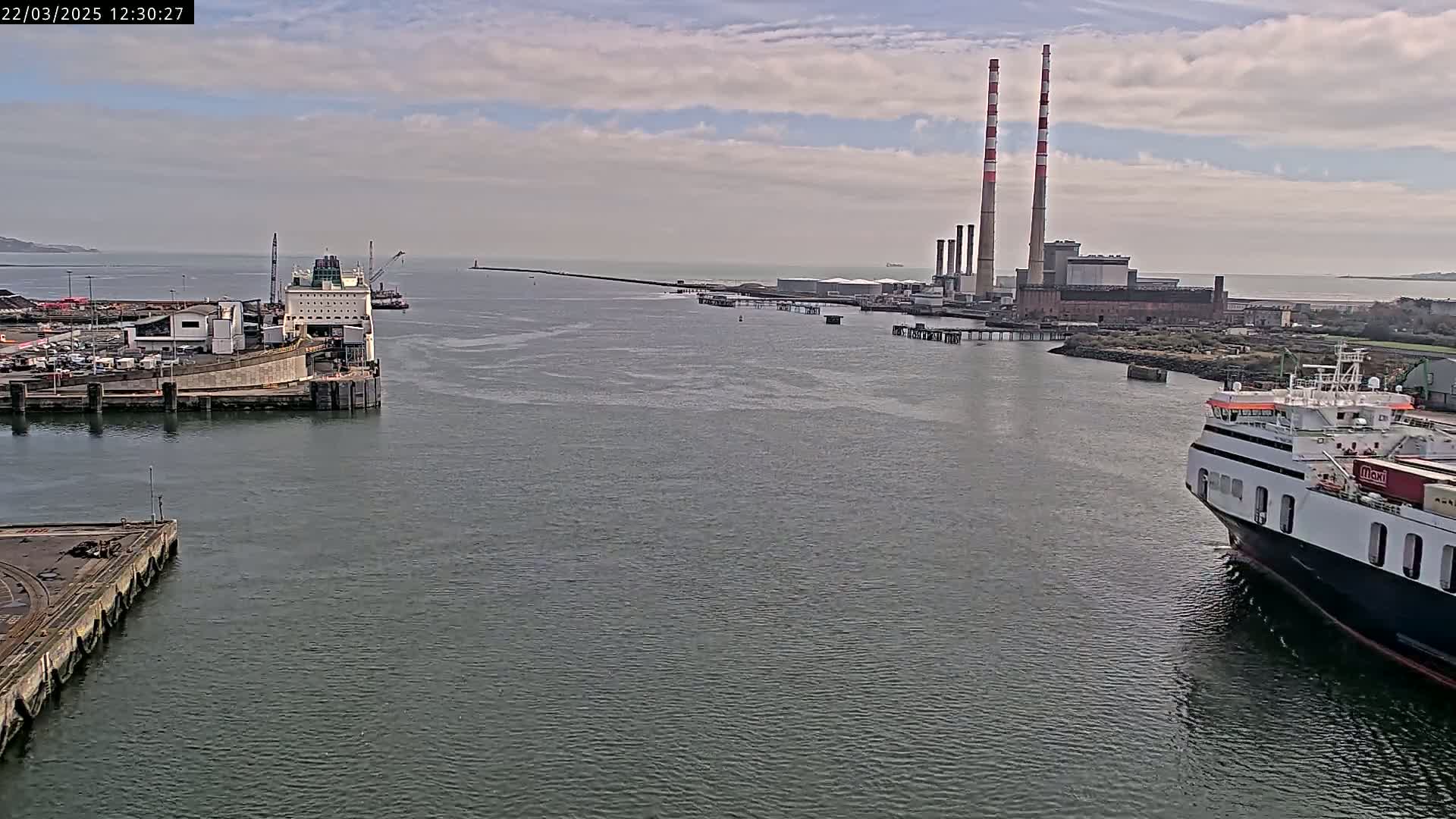A harbor scene under a partly cloudy sky shows two ferries docked near a power plant with tall smokestacks.