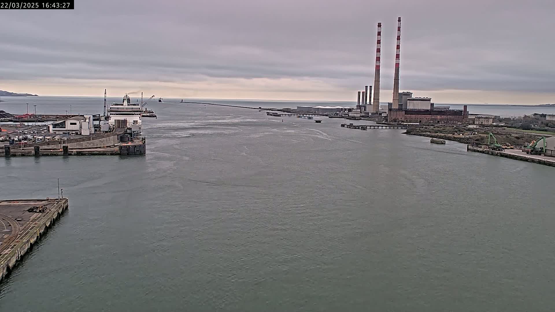 A ferry is docked at a harbor near a power plant with two tall smokestacks under an overcast sky.