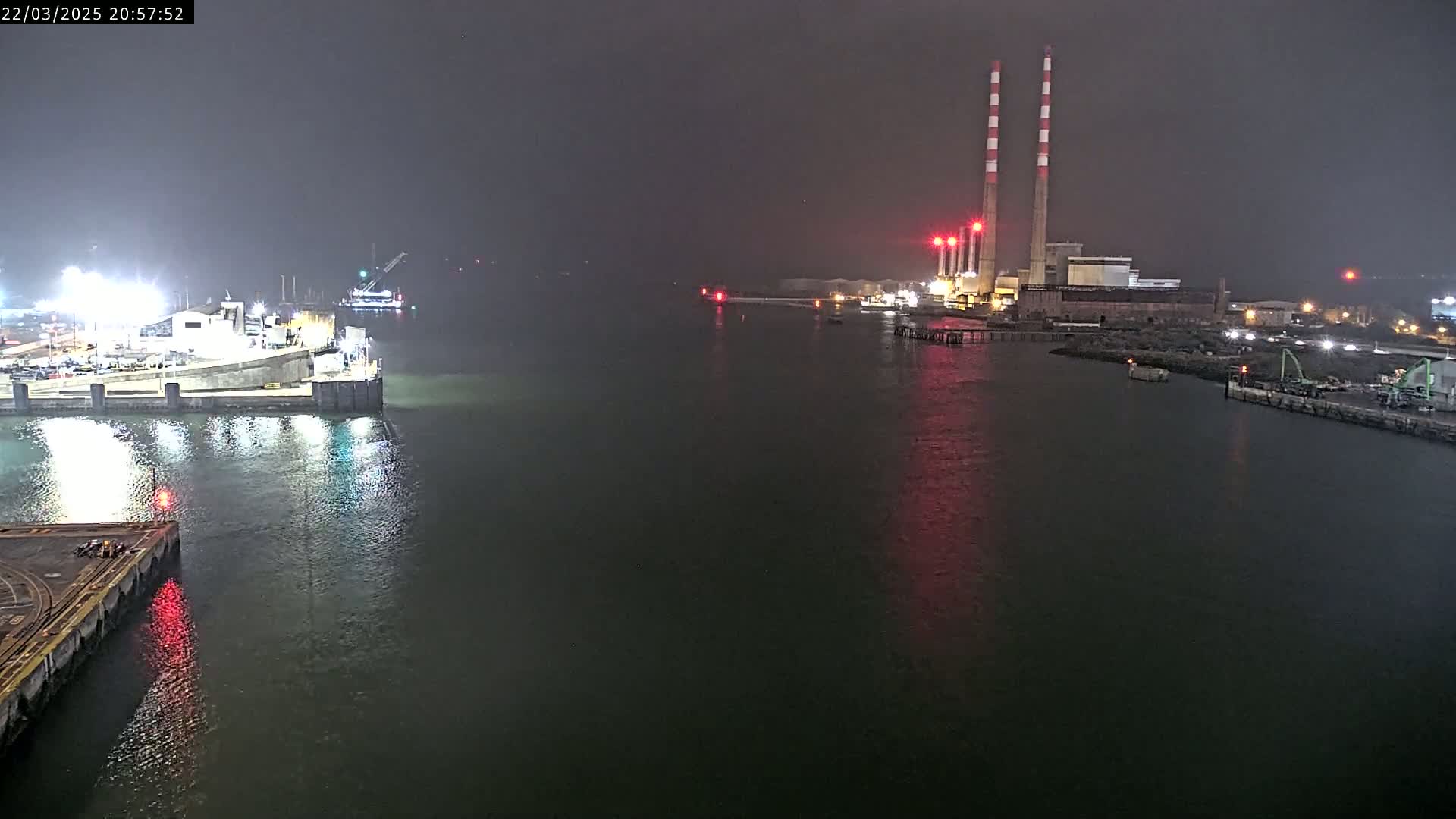 A nighttime view of a harbor shows a dock with lights, a body of water, and a power plant with tall smokestacks in the distance under foggy conditions.