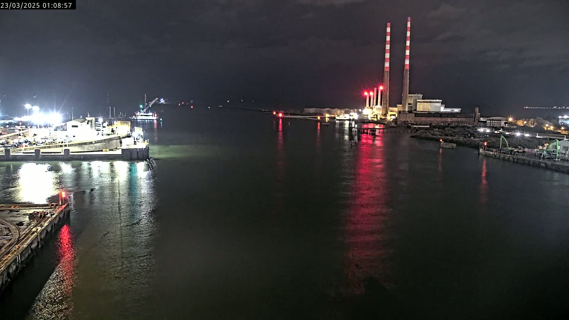 A nighttime view of a calm body of water with industrial structures and docked vessels, lit by artificial lights.