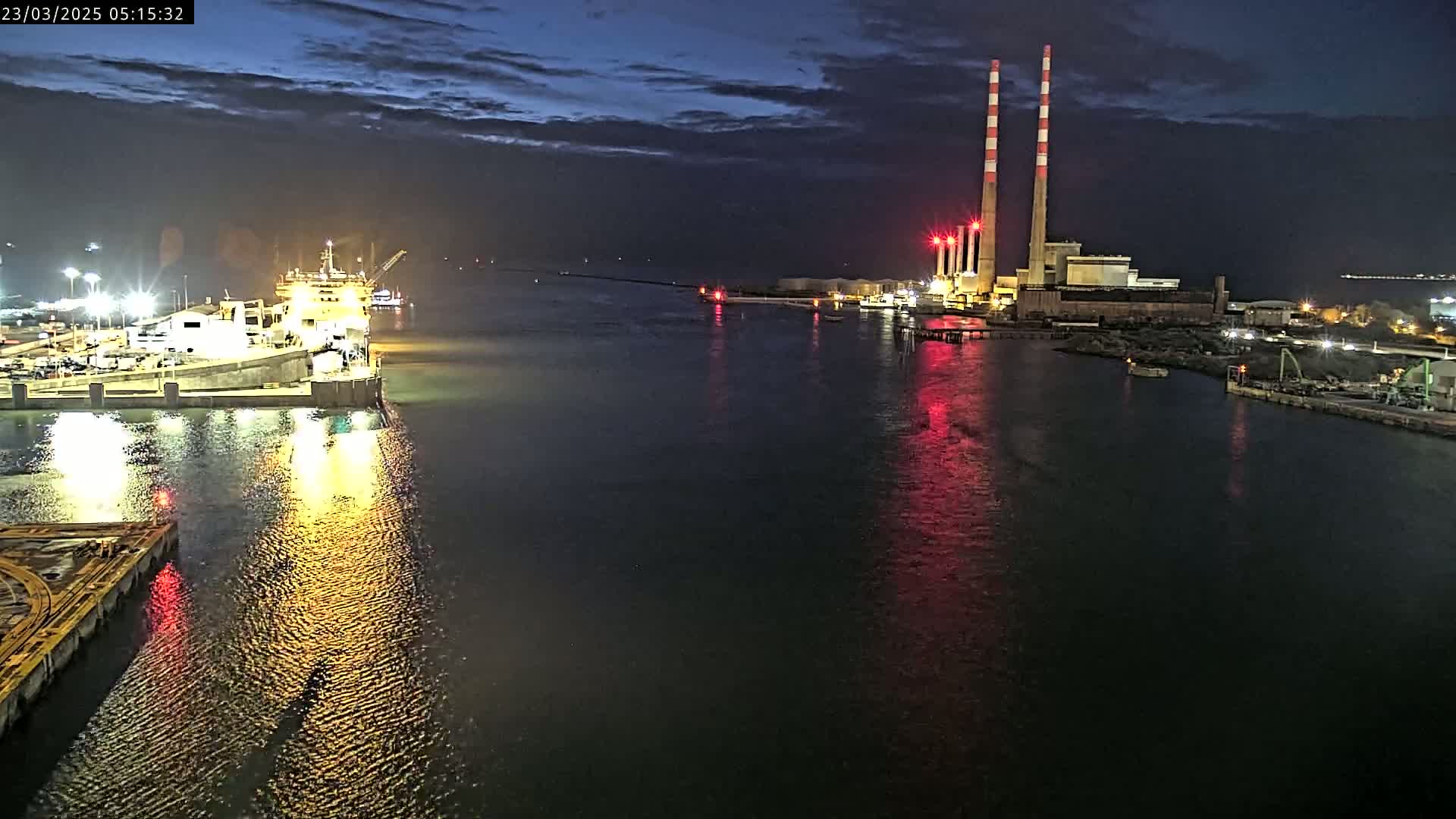A large ship is docked at a brightly lit port at night under a partly cloudy sky, with a power plant and its tall red and white smokestacks visible in the distance across the water.