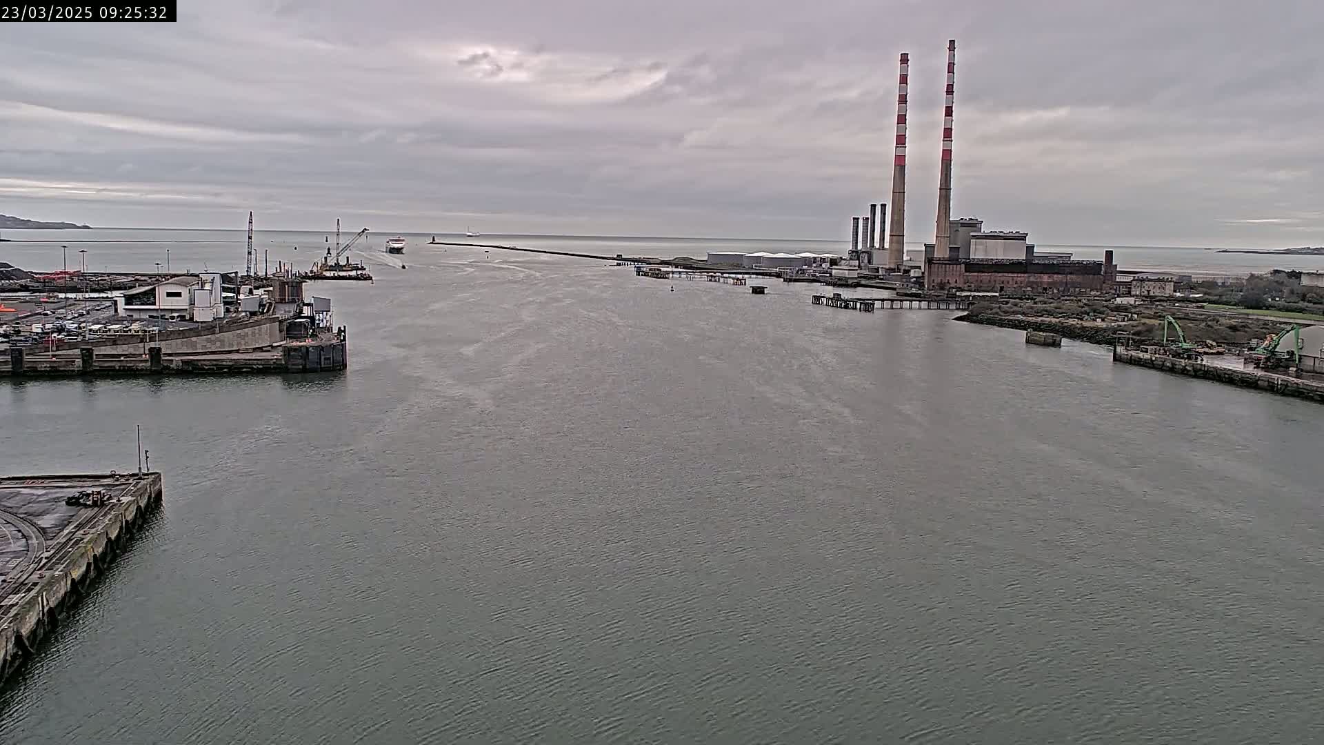 A wide view of a harbor on an overcast day shows a large industrial facility with tall smokestacks, a ship in the distance, and various docks and structures along the waterway.