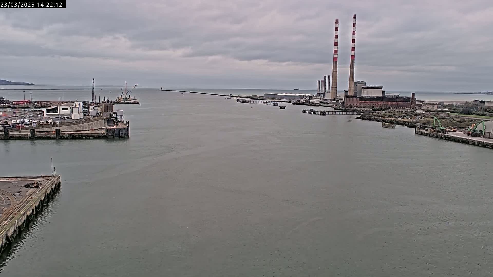 A wide shot of a harbor on an overcast day, showing a long pier extending into the water toward a power plant with tall smokestacks.