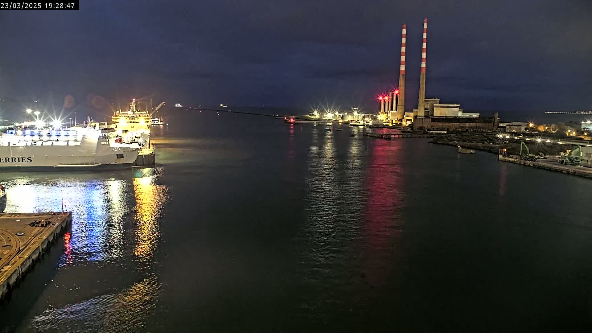 A ferry is docked at night next to a brightly lit industrial facility with two tall smokestacks, reflected in the dark water.