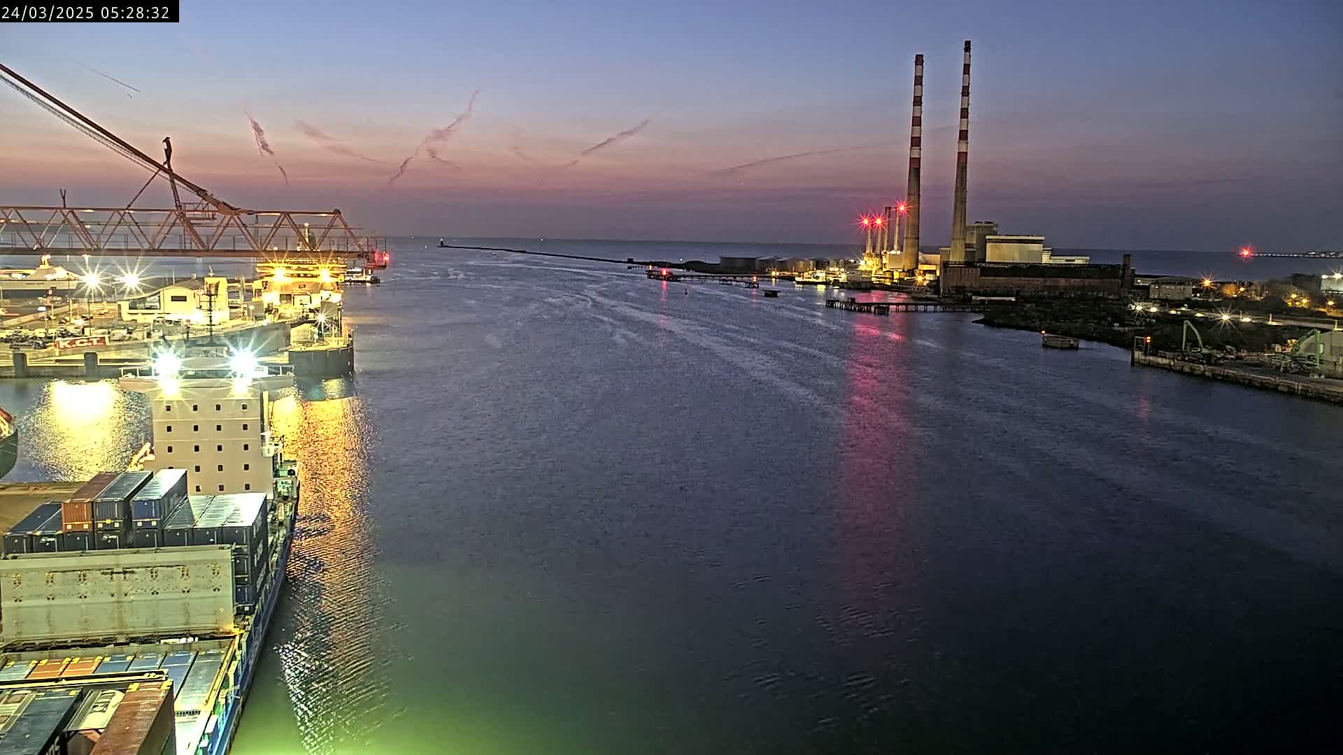A container ship is docked at a port at twilight, with a power plant visible in the background under a mostly clear sky.