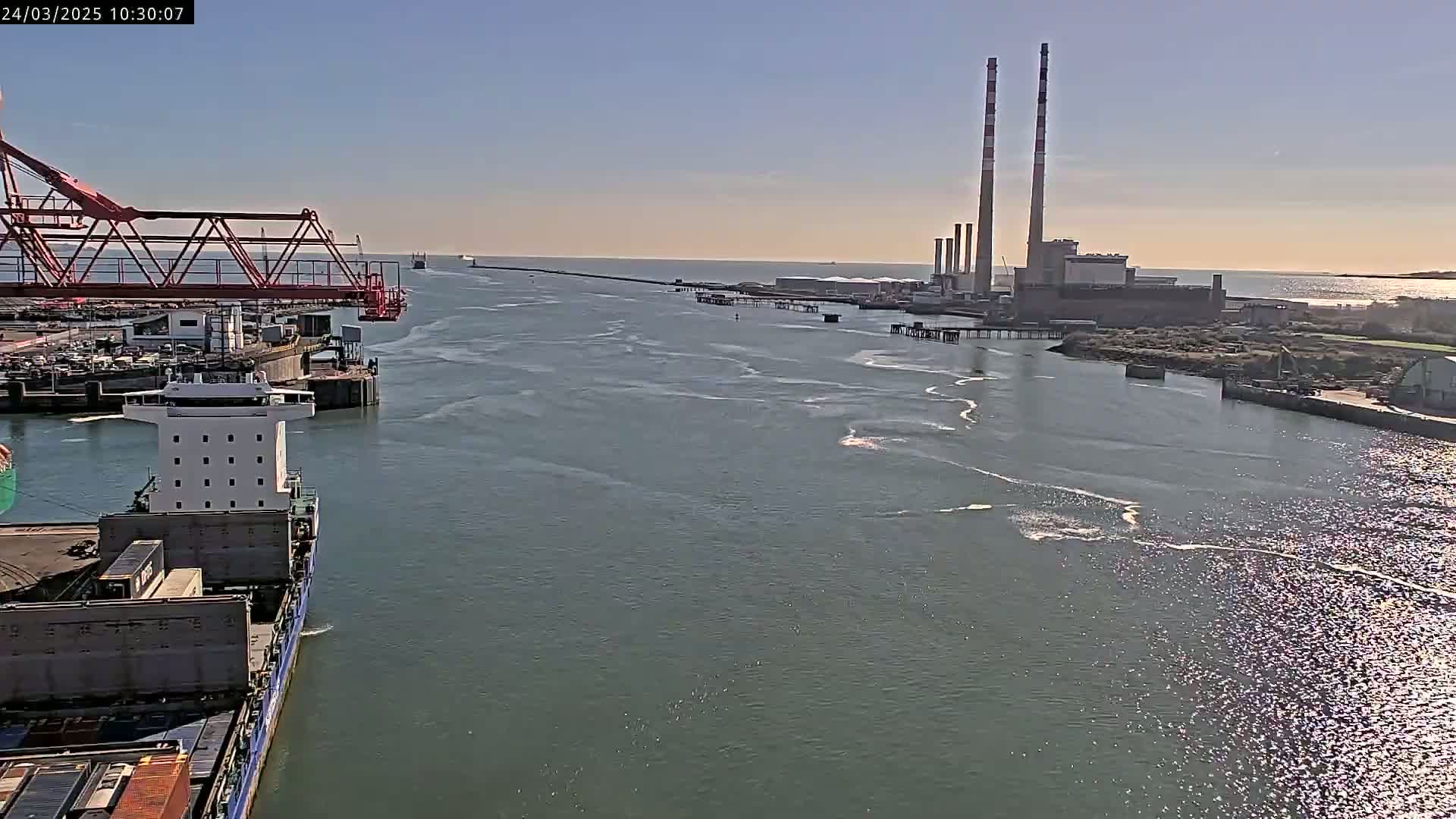 A large cargo ship is seen leaving a port under sunny conditions, with a power plant and a long jetty visible in the distance.