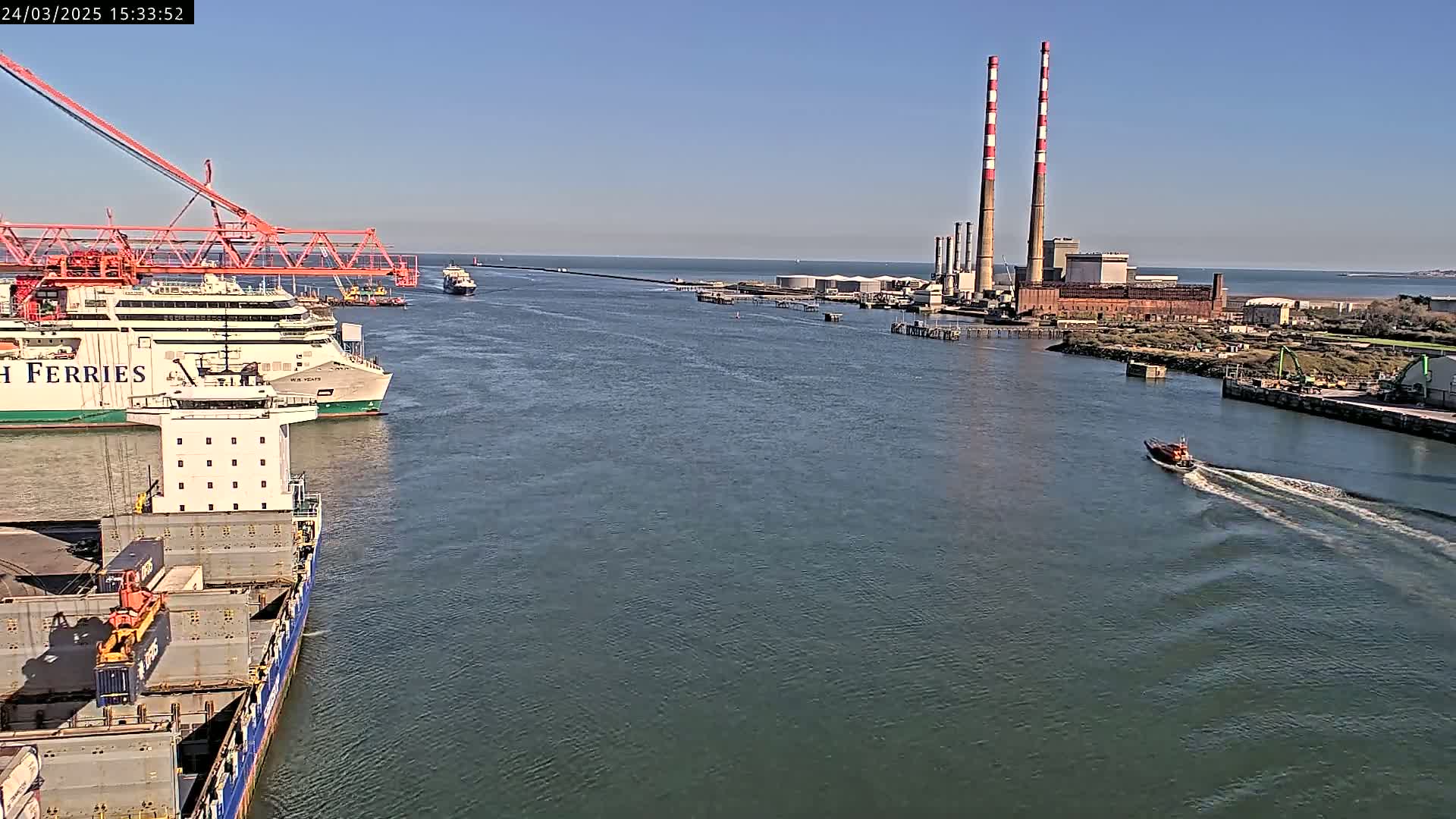 A ferry and cargo ship are in a harbor on a sunny day, with a power plant visible in the background.