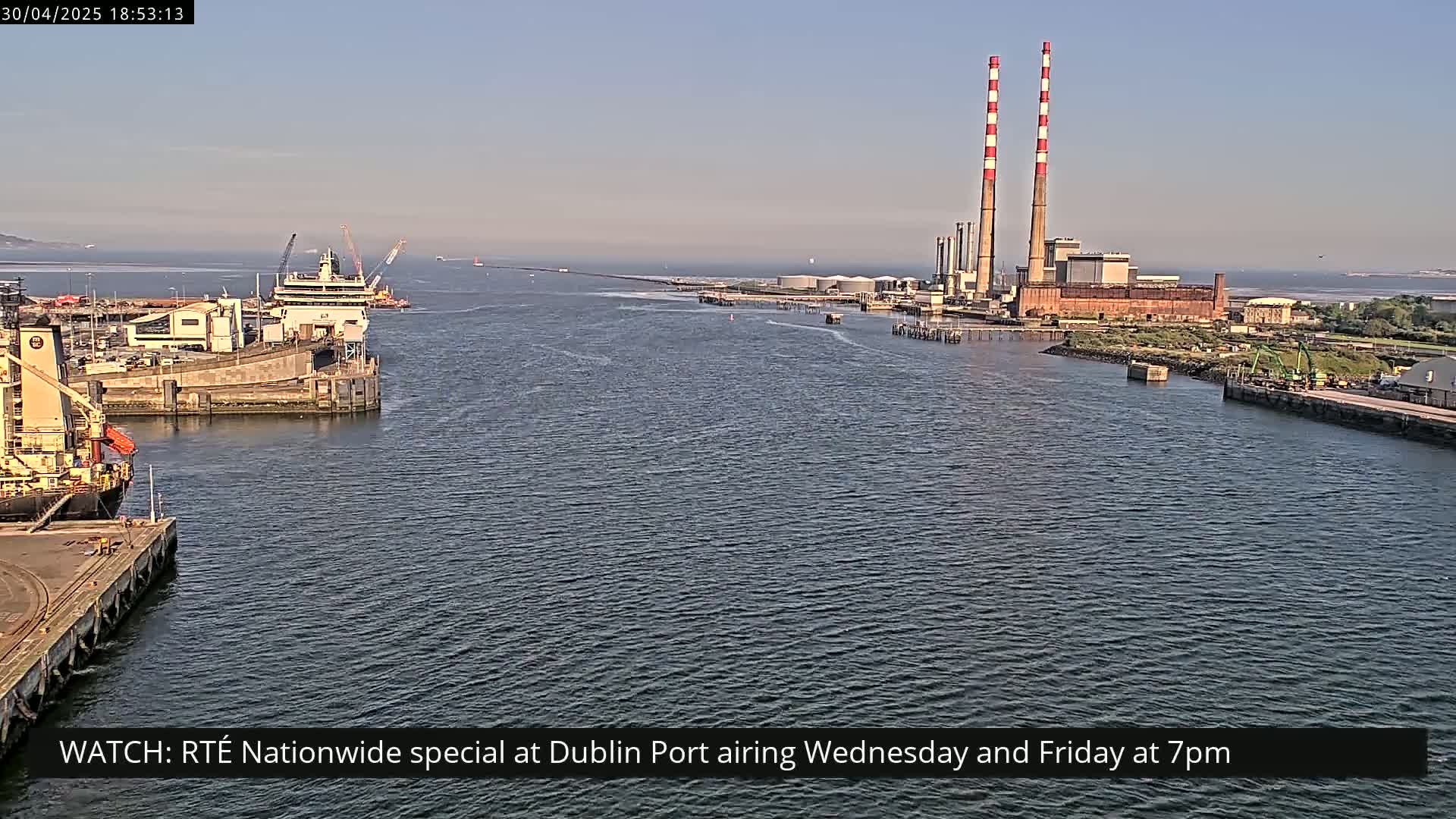 A calm harbor scene shows ships docked near a large industrial plant with tall smokestacks under a clear, sunny sky.