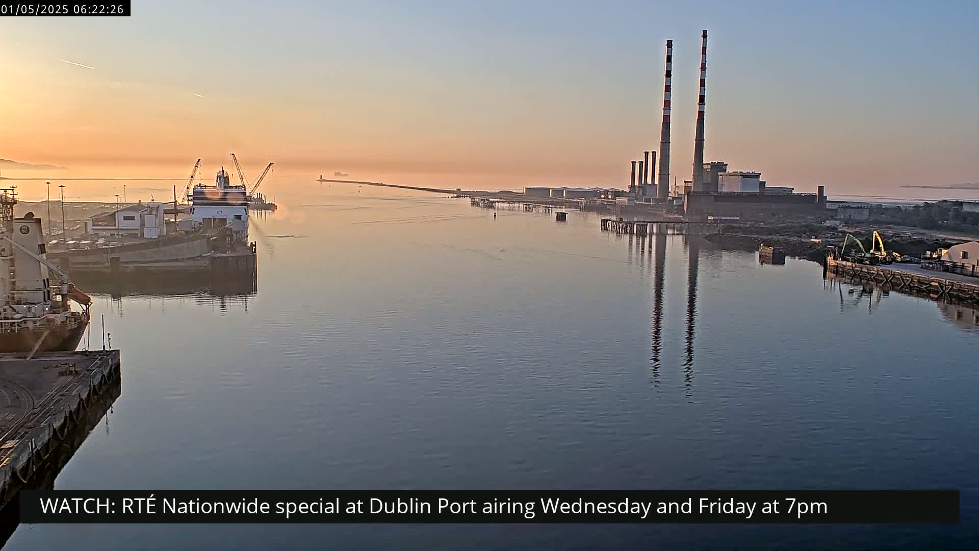 A calm body of water at sunrise shows ships docked at a port with two large industrial chimneys in the background and a light fog covering the horizon.