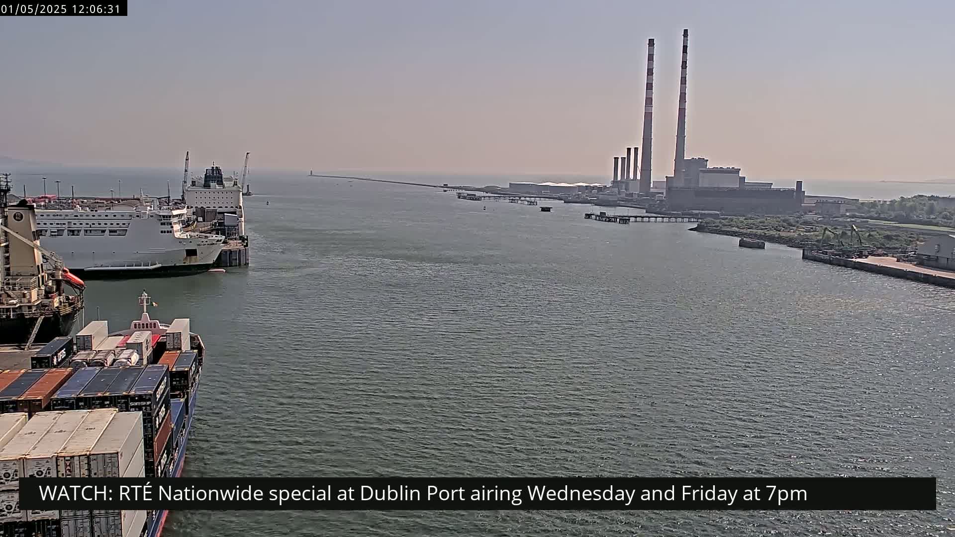 A container ship and a ferry are docked in a harbor, with a power plant visible in the background on a hazy day.