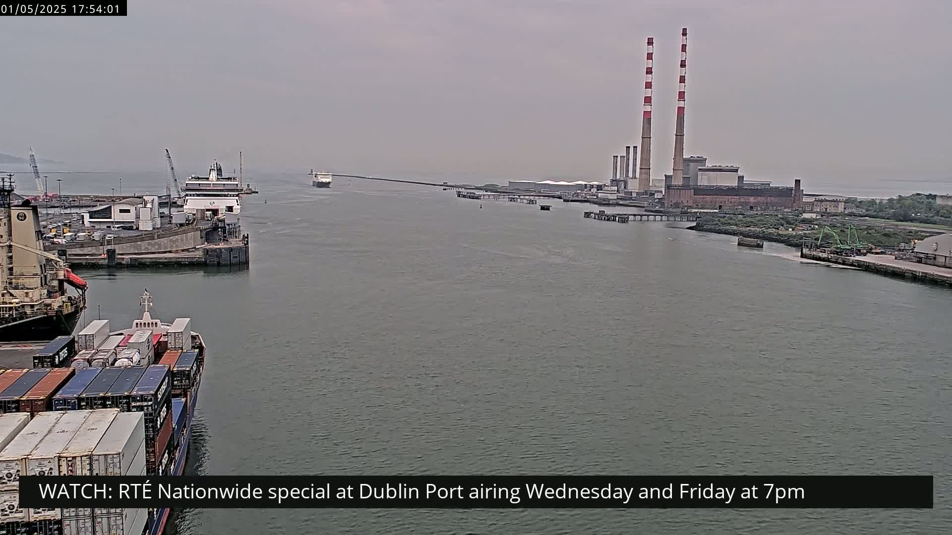 A container ship and a passenger ferry are docked at a port under an overcast sky, with tall industrial smokestacks visible in the distance.