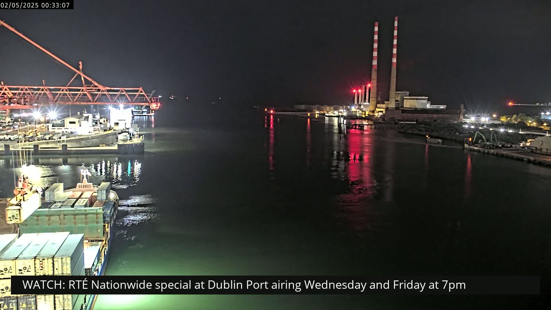 At night, a dark body of water reflects the lights of a port with a large industrial structure and several ships.