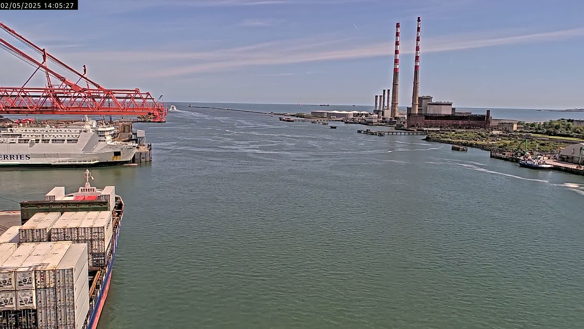 A container ship and ferry are docked near a power plant with tall smokestacks on a sunny day.