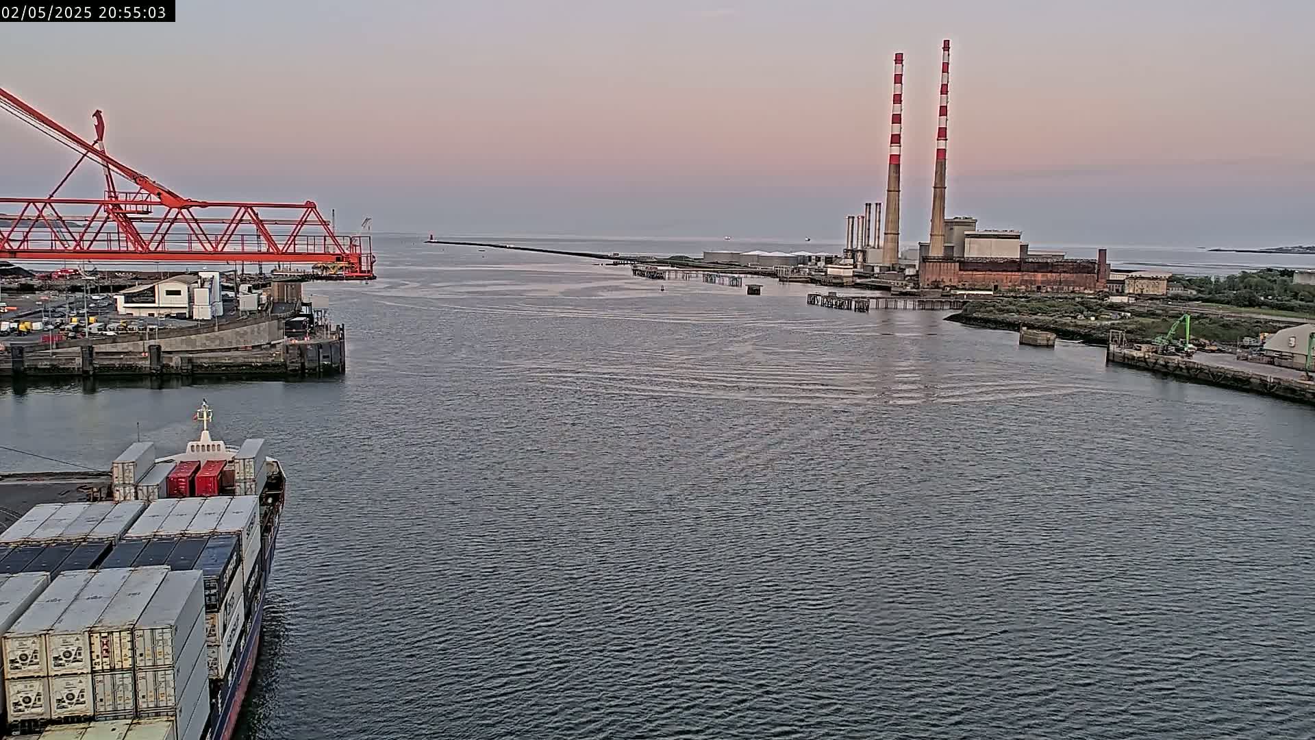 A container ship sails past a large industrial plant with tall smokestacks under a dusky sky.