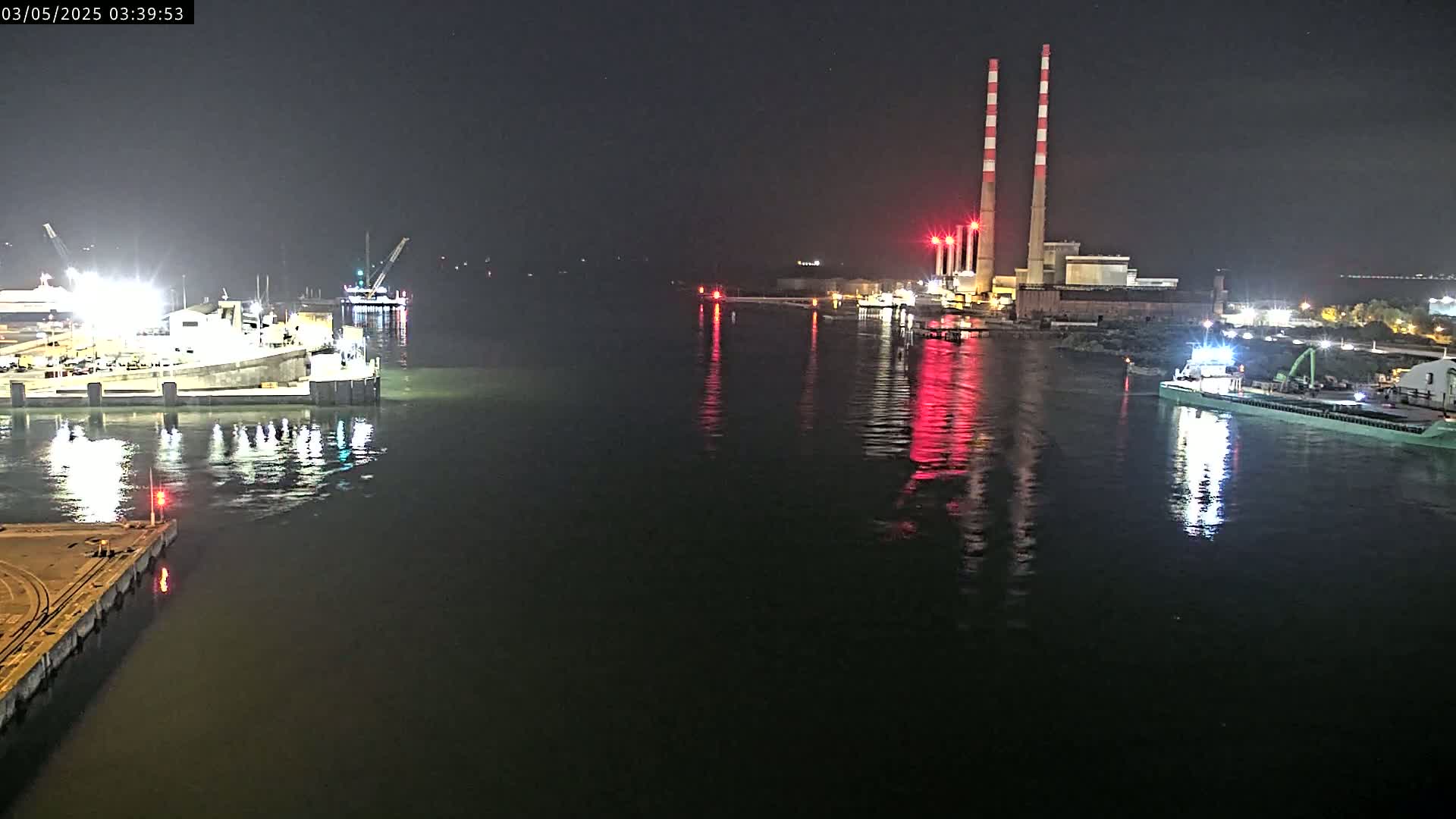 At night, a harbor scene shows several docked ships and boats near a power plant with tall red and white smokestacks reflected in calm dark water.