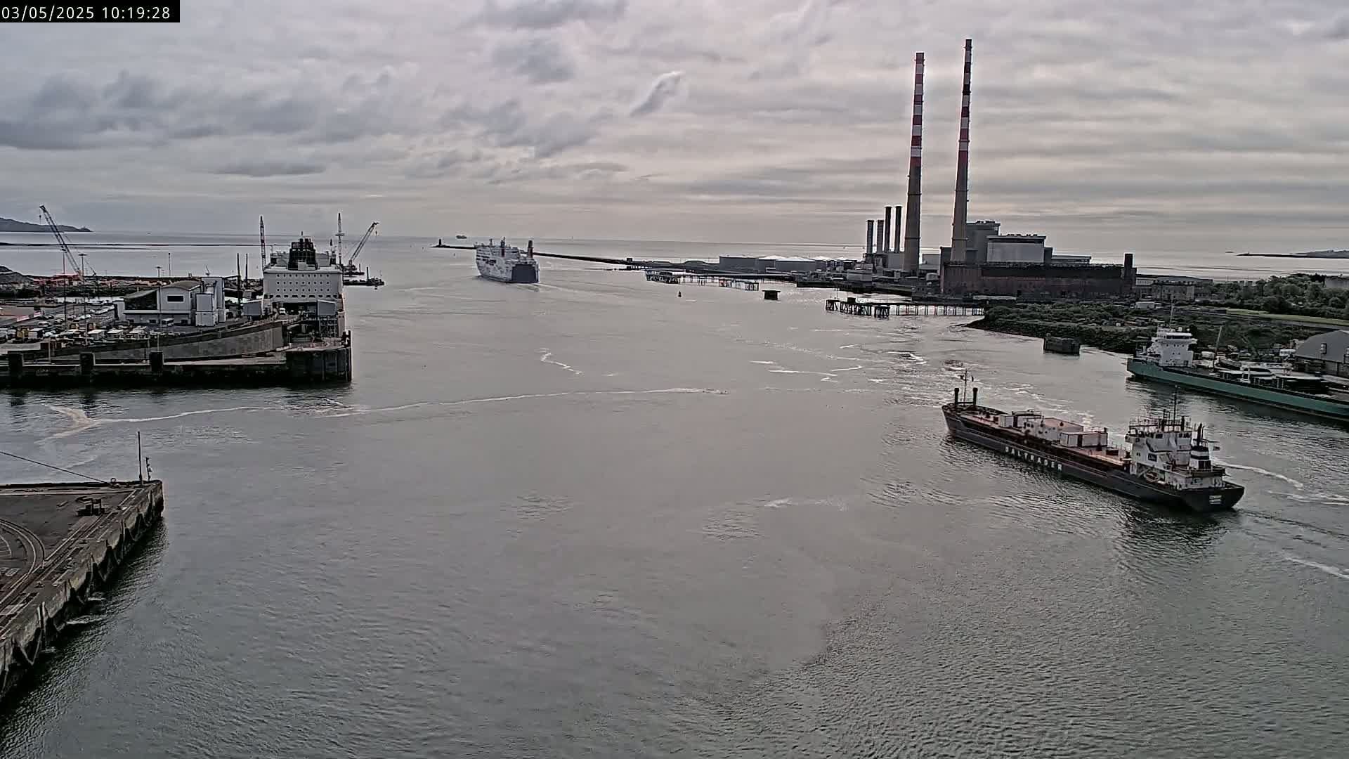 Several ships are navigating a harbor under a cloudy sky, with a power plant visible in the background.