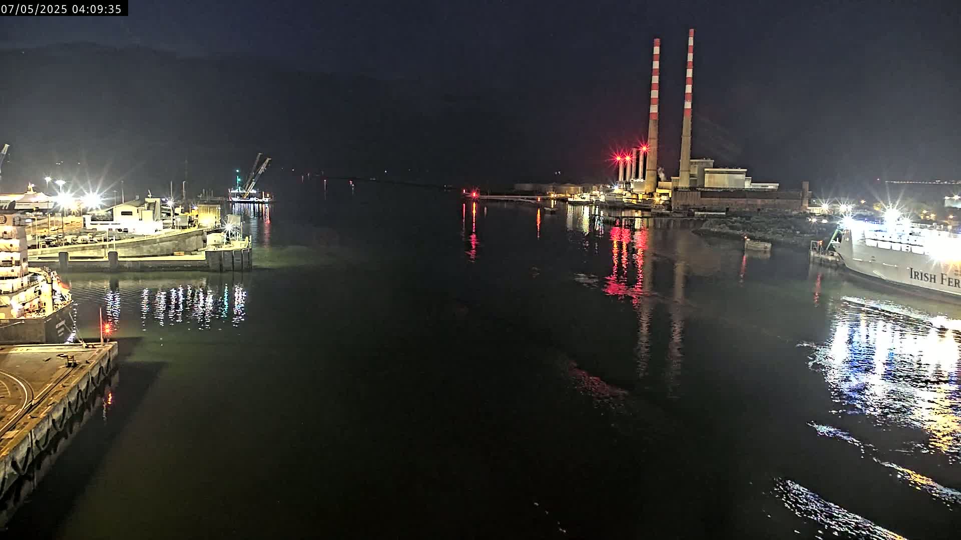 At night, a large ferry is docked near a power plant with tall smokestacks, while other ships are moored at a nearby dock in calm water.