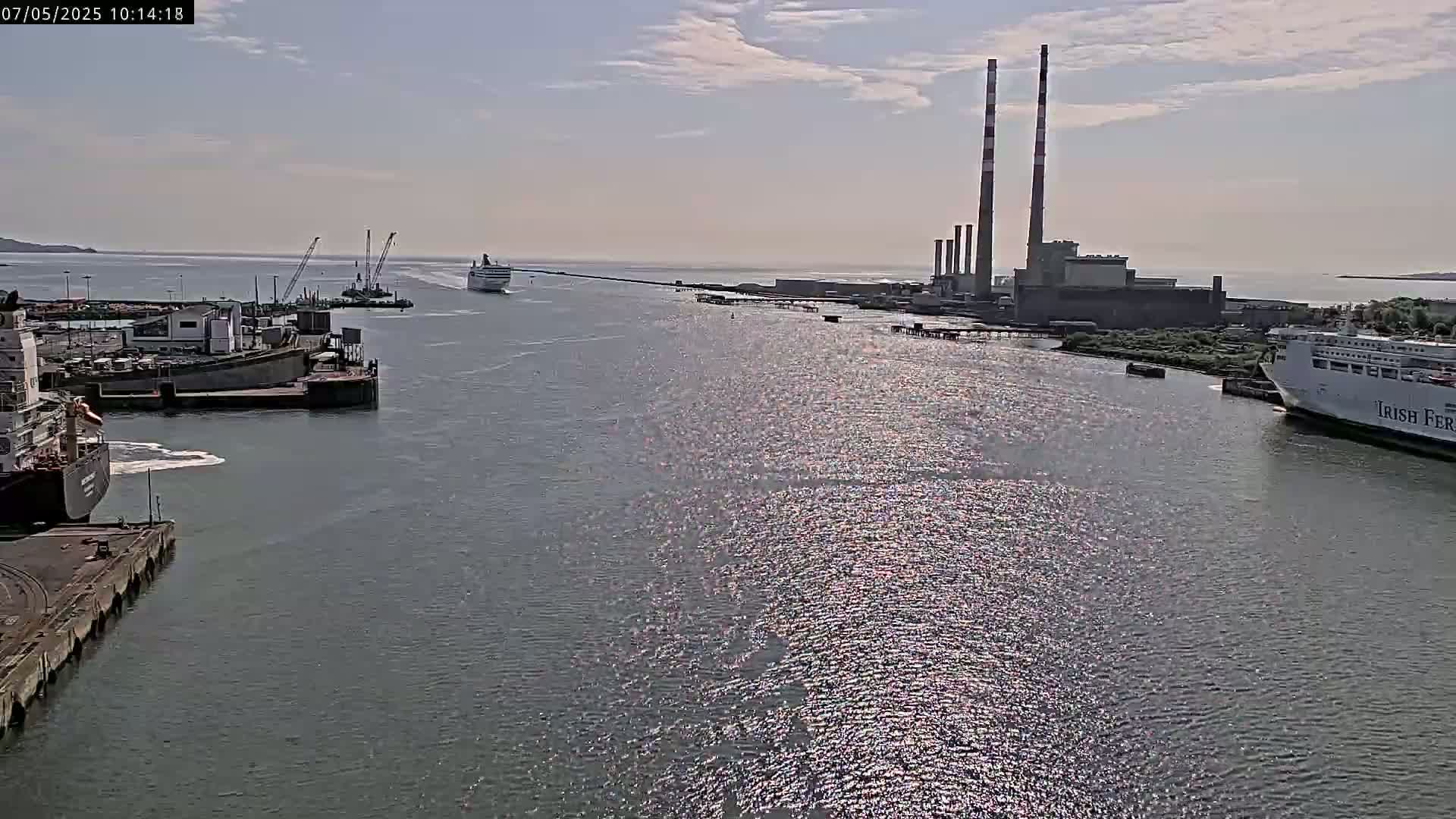 A wide harbor scene on a sunny day shows several ships, including a large ferry, docked and underway near a power plant with tall smokestacks.
