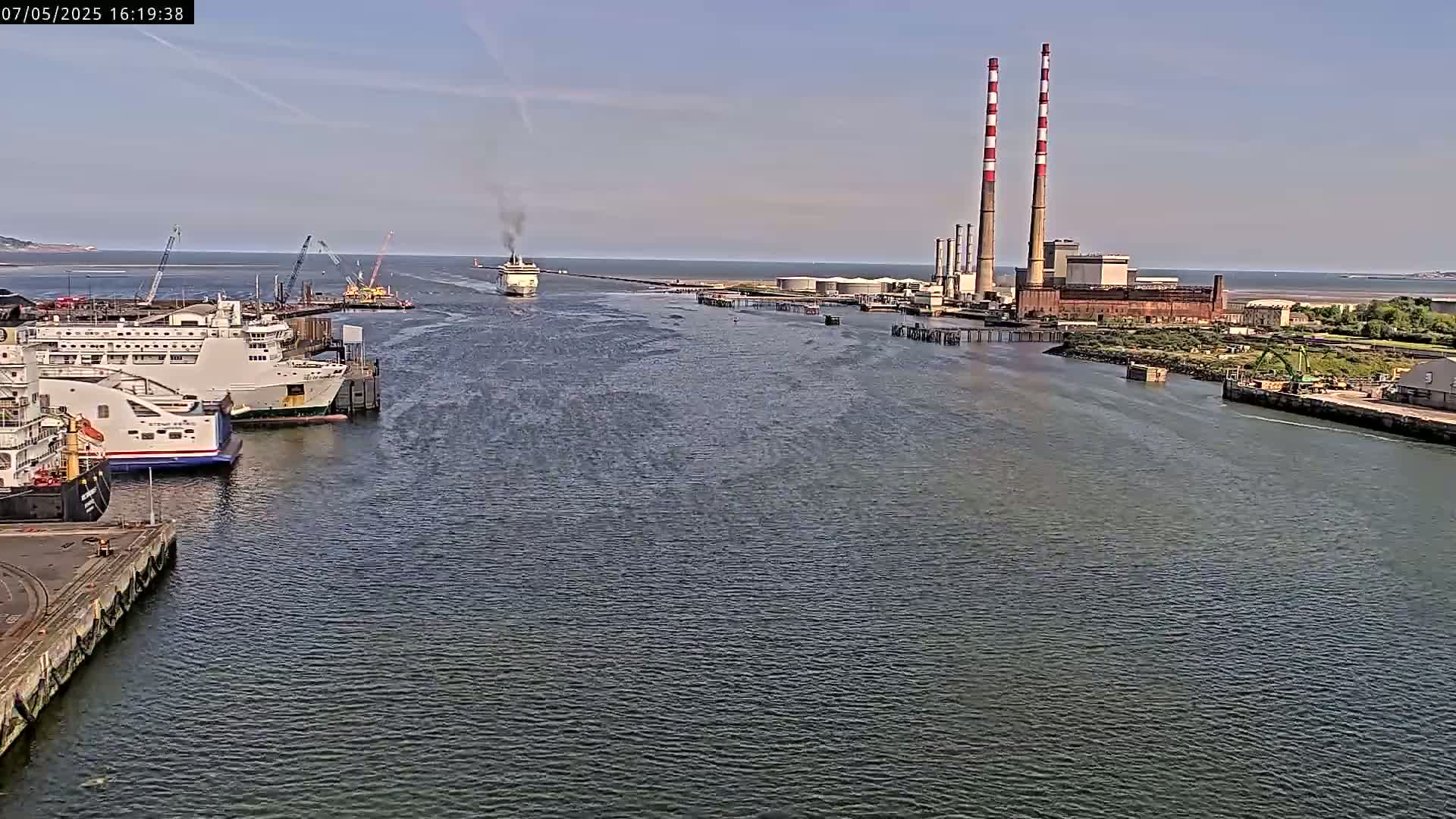 A ferry leaves a harbor on a calm, sunny day, passing by docked ships and a power plant with tall smokestacks in the background.