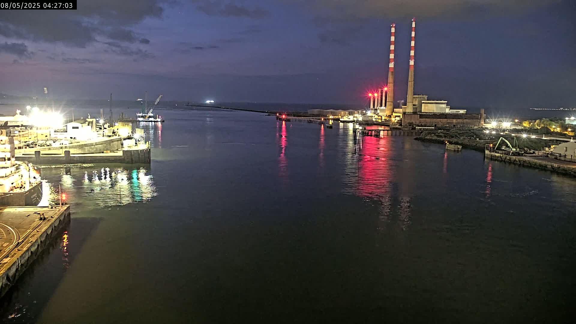 A nighttime view of a calm harbor with docked ships, a power plant with tall smokestacks, and the reflections of lights on the water.