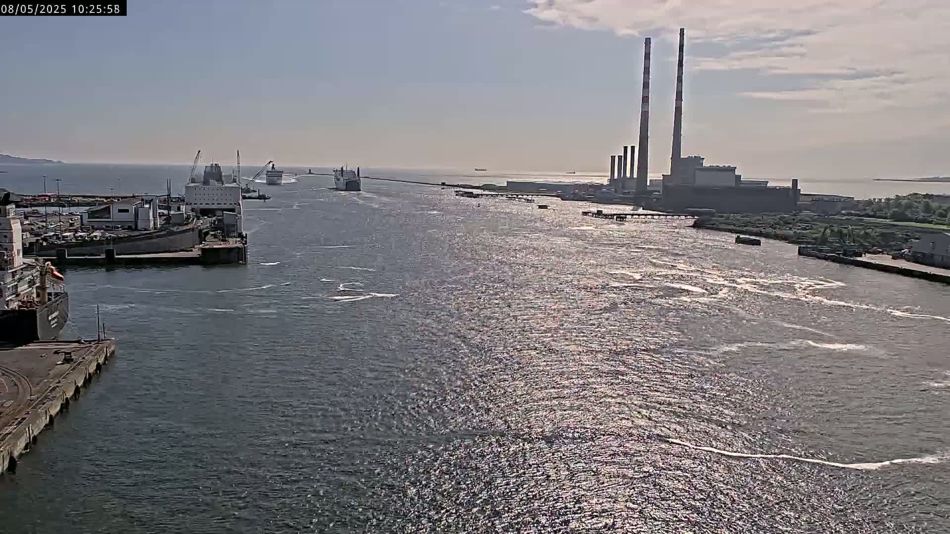 Several ships are docked and sailing in a harbor on a partly sunny day, with a large industrial facility visible in the distance.
