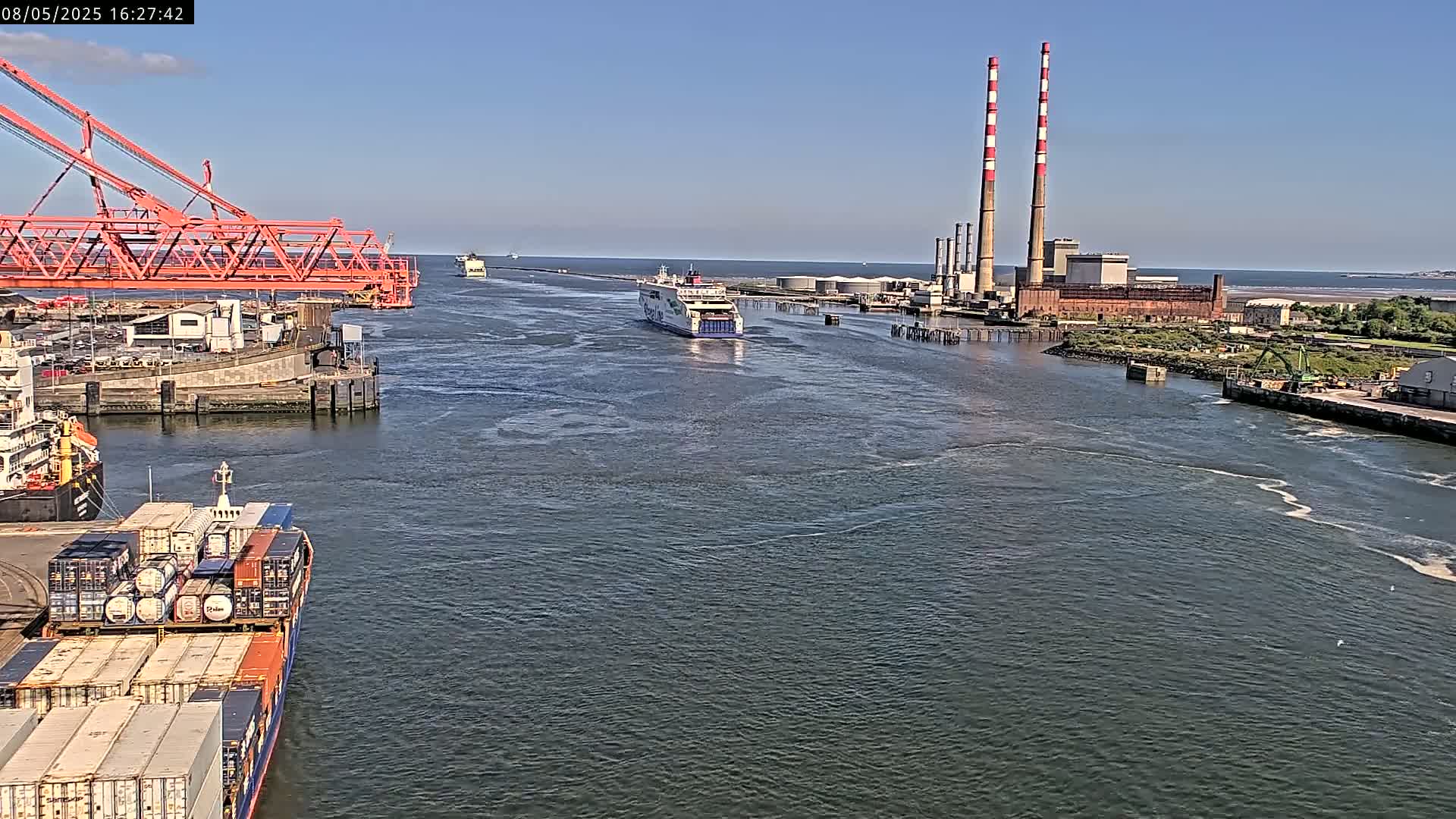 A ferry passes a container ship and industrial plant with tall chimneys on a sunny day in a harbor.
