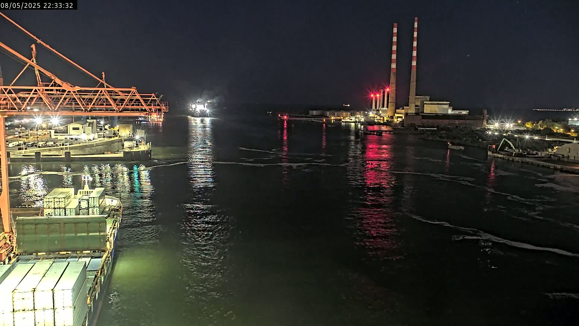 At night, a container ship is docked at a port near a power plant with two tall smokestacks, under a clear, dark sky.